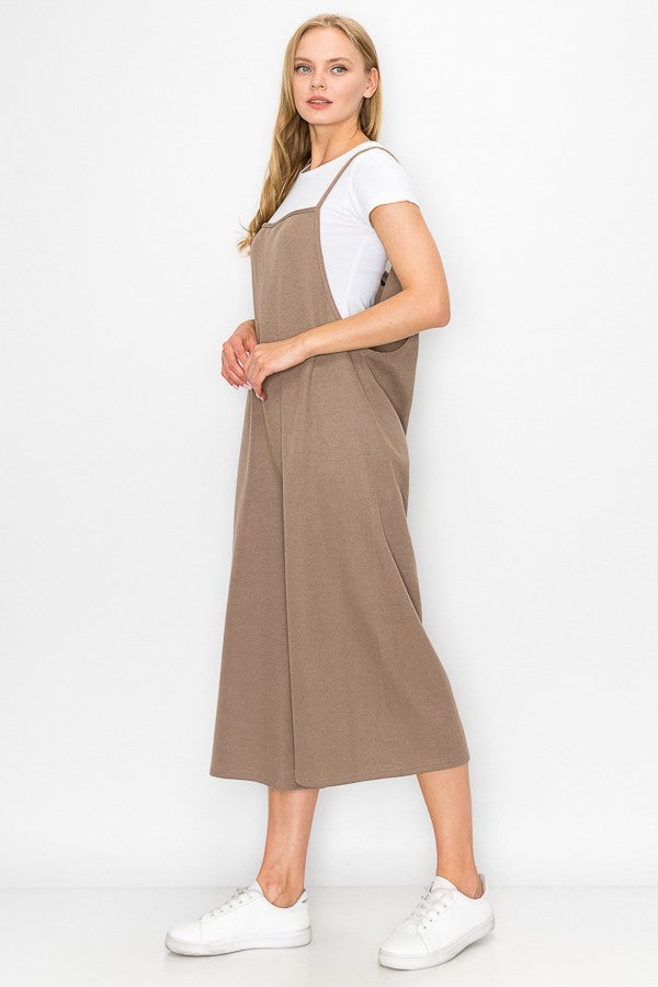 A person with long blonde hair sits on a wooden stool against a plain background, wearing Stylespects Cropped Bottom Wide Leg Oversized Jumpsuit in taupe and white sneakers.