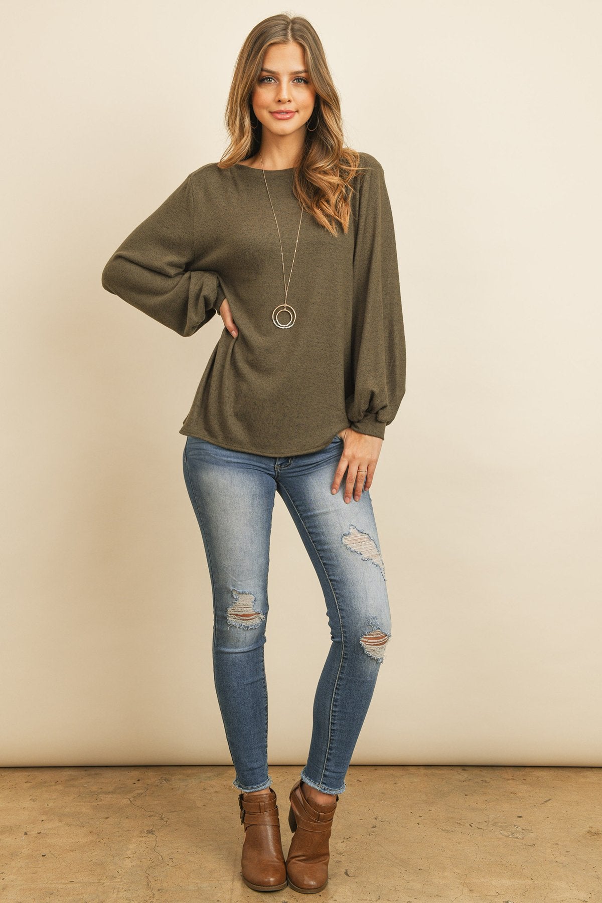 A woman models a Riah Fashion Puff Sleeved Boat Neck Two Toned Brushed Hacci Top with ripped jeans and brown boots on a concrete floor against a plain backdrop, accessorized with a long necklace.