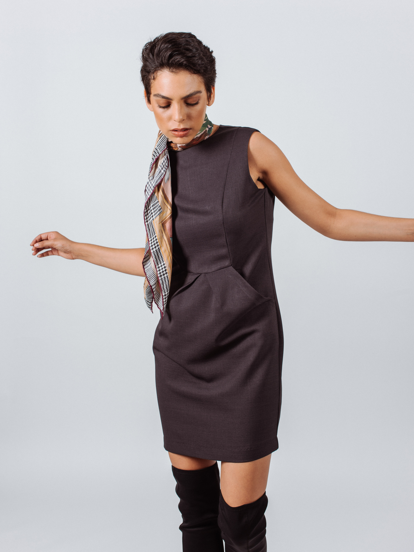 A woman in a sleeveless black Le Réussi® Italian Wool Dress Pleated With Pockets and black heels stands with hands on hips, facing forward against a white background.