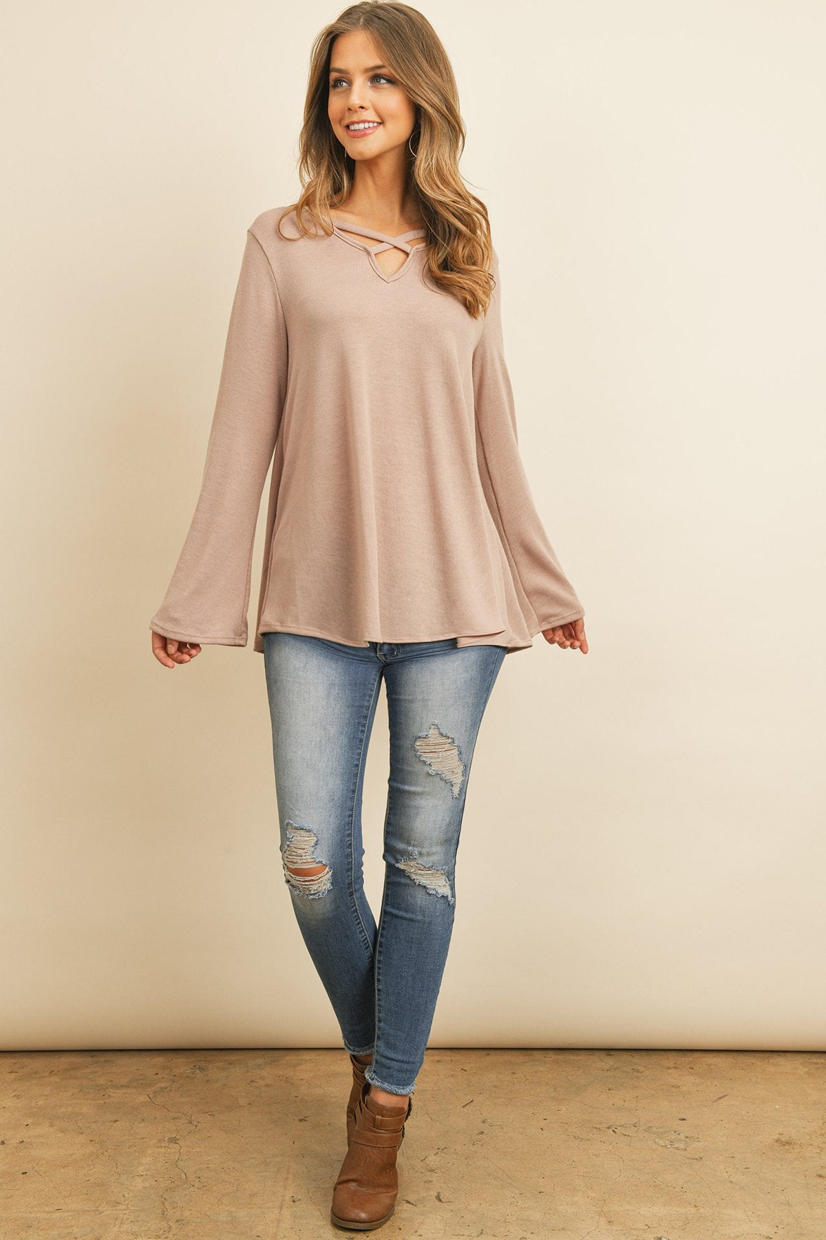 A person wearing a Riah Fashion Criss Cross Neck Long Sleeve Hacci Top in Dusty Blush and blue jeans stands against a neutral background, smiling.