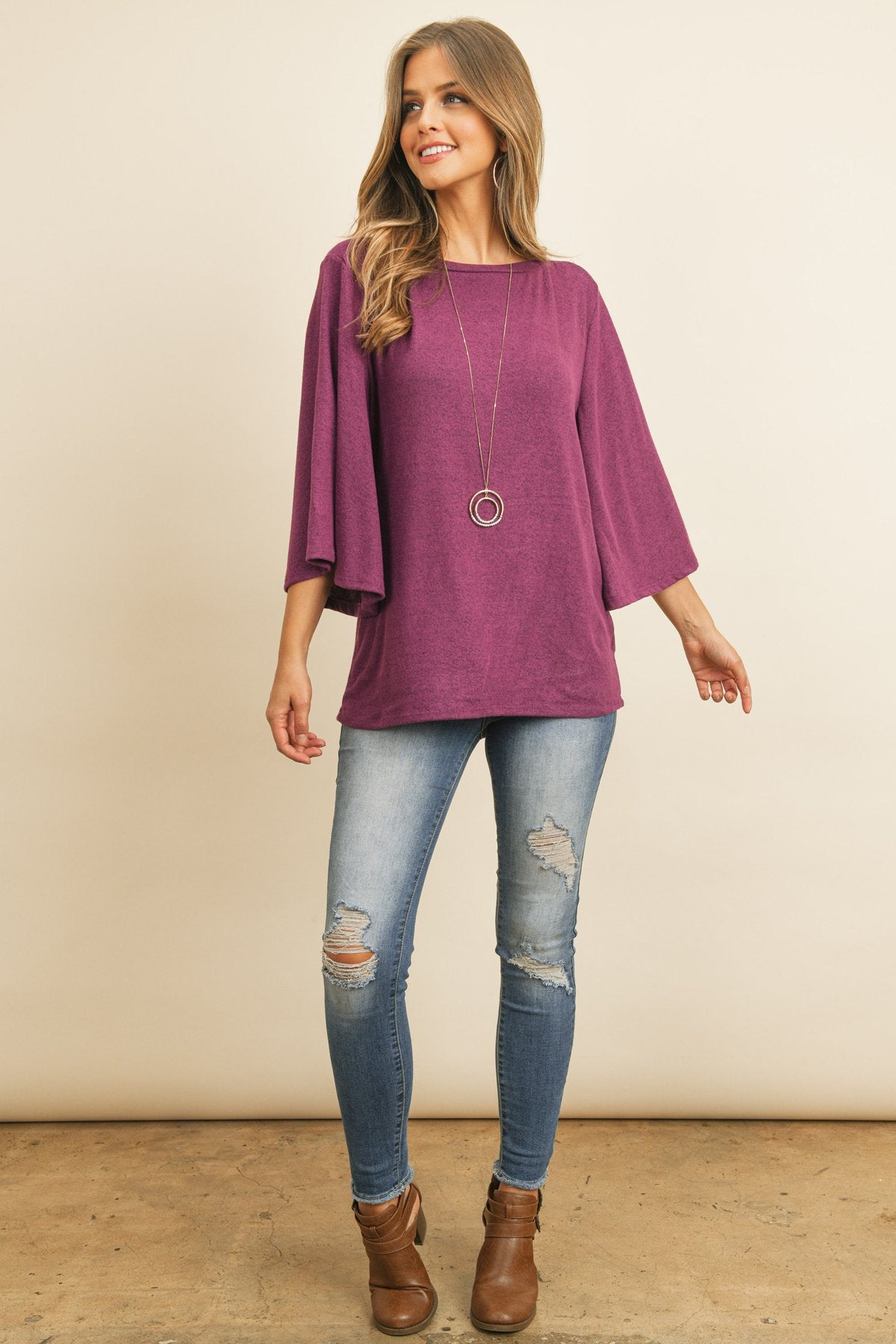 A person in a Riah Fashion Boat Neck Bell Sleeve Solid Hacci Brushed Top and ripped jeans stands against a neutral background, accessorized with hoop earrings and a long necklace.