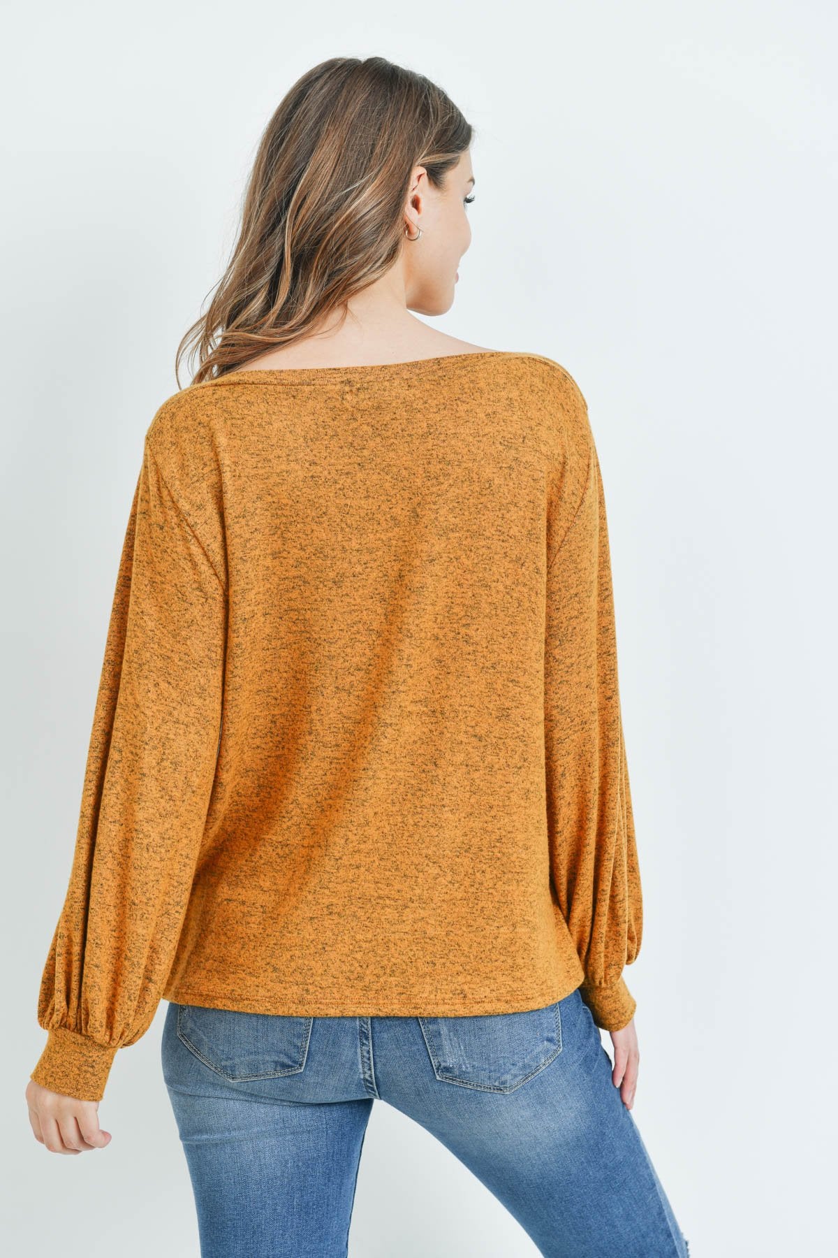 A woman models a Riah Fashion Puff Sleeved Boat Neck Two Toned Brushed Hacci Top with ripped jeans and brown boots on a concrete floor against a plain backdrop, accessorized with a long necklace.
