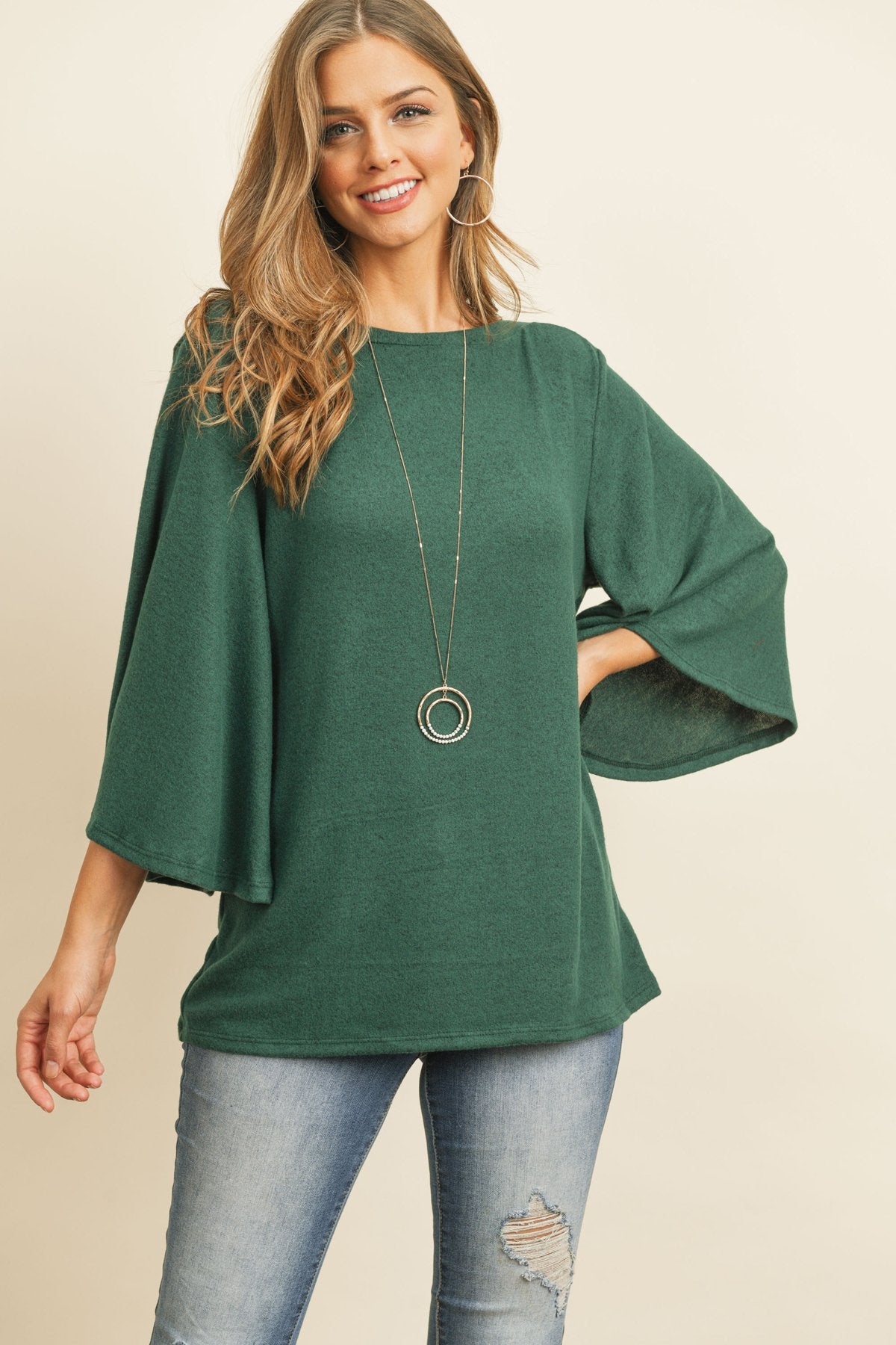 A person in a Riah Fashion Boat Neck Bell Sleeve Solid Hacci Brushed Top and ripped jeans stands against a neutral background, accessorized with hoop earrings and a long necklace.