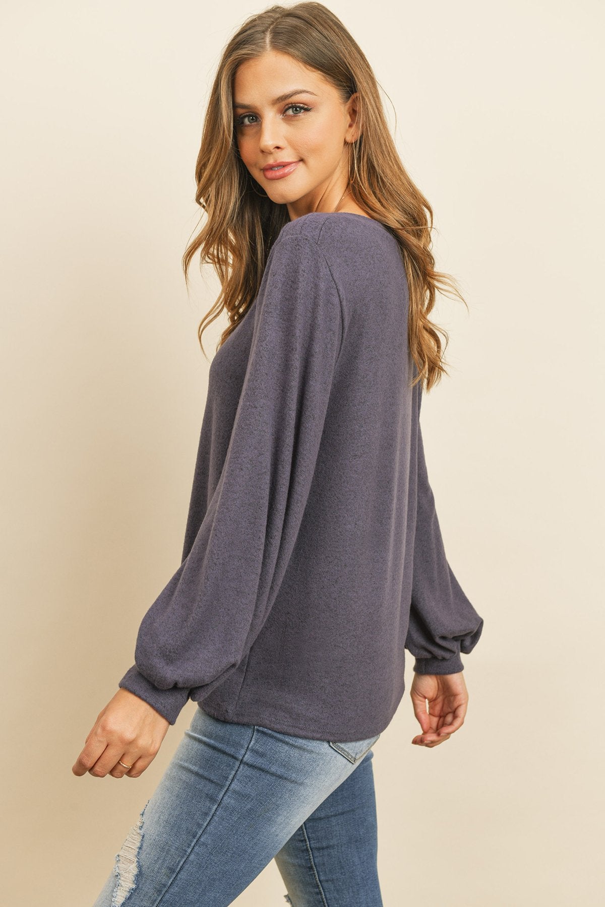 A woman models a Riah Fashion Puff Sleeved Boat Neck Two Toned Brushed Hacci Top with ripped jeans and brown boots on a concrete floor against a plain backdrop, accessorized with a long necklace.