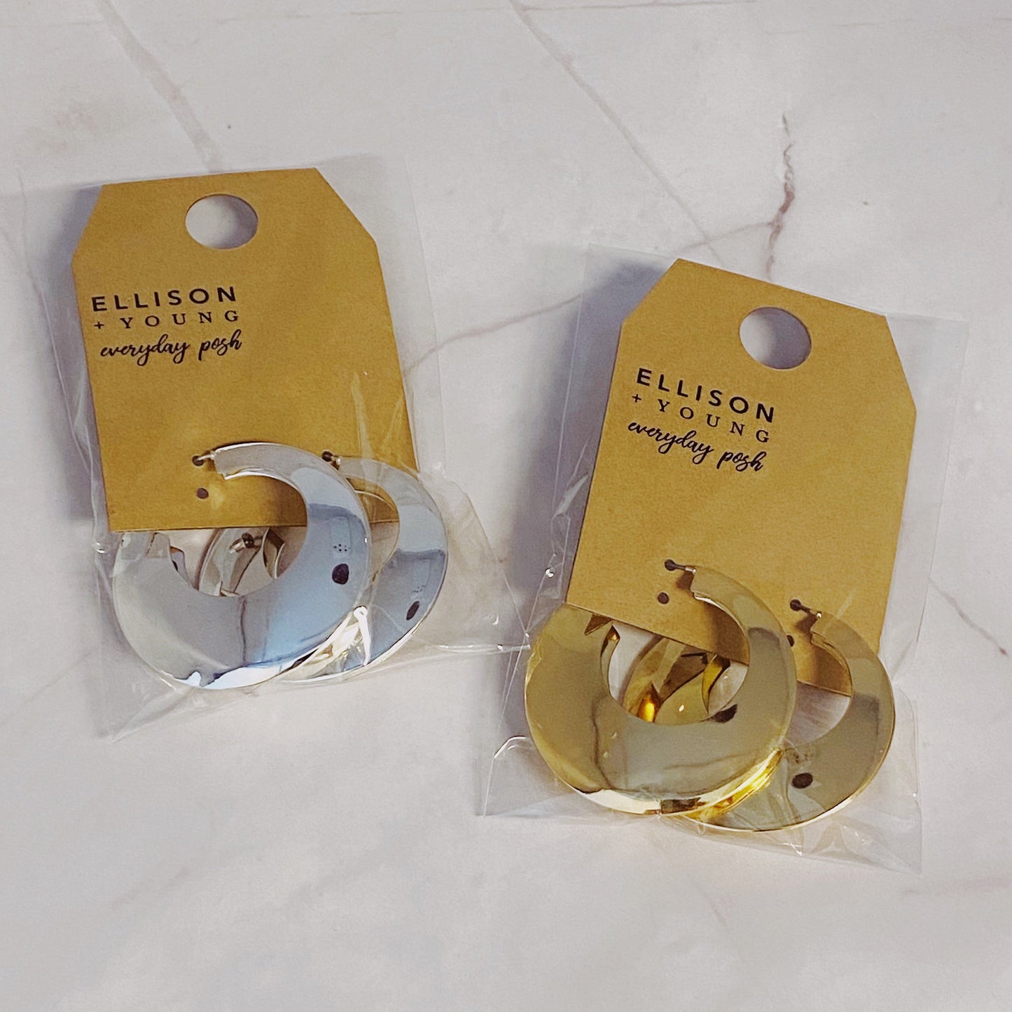 On a marble surface, four round Light and Modern Hoop Earrings from Ellison + Young are displayed, with two in silver and two in gold.