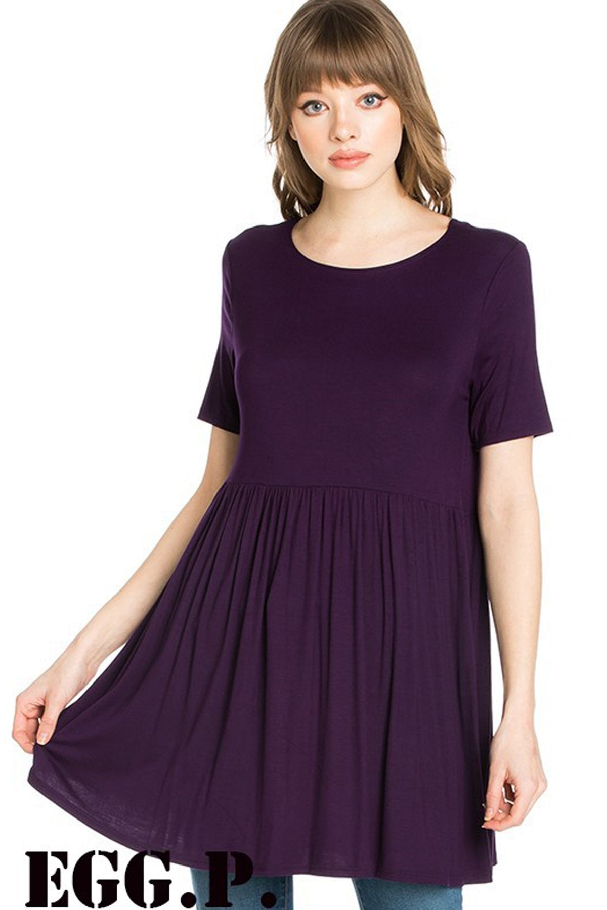 A woman stands outdoors wearing a beige Riah Fashion Short Sleeve Empire Waist Tunic paired with a long necklace.