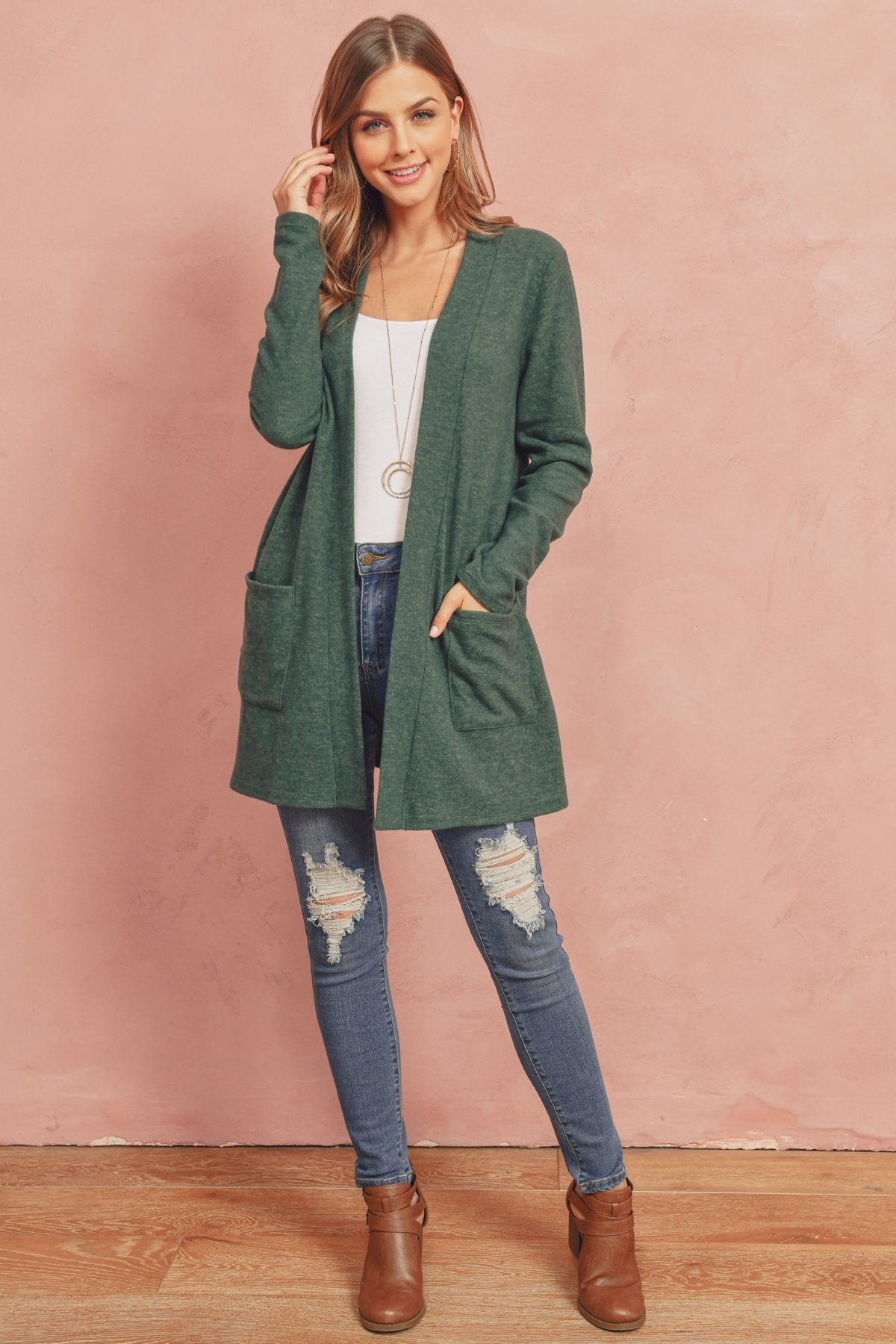 A woman in a Brushed Melange Open Front Cardigan by Riah Fashion and ripped jeans stands against a pink background, hands in her pockets.