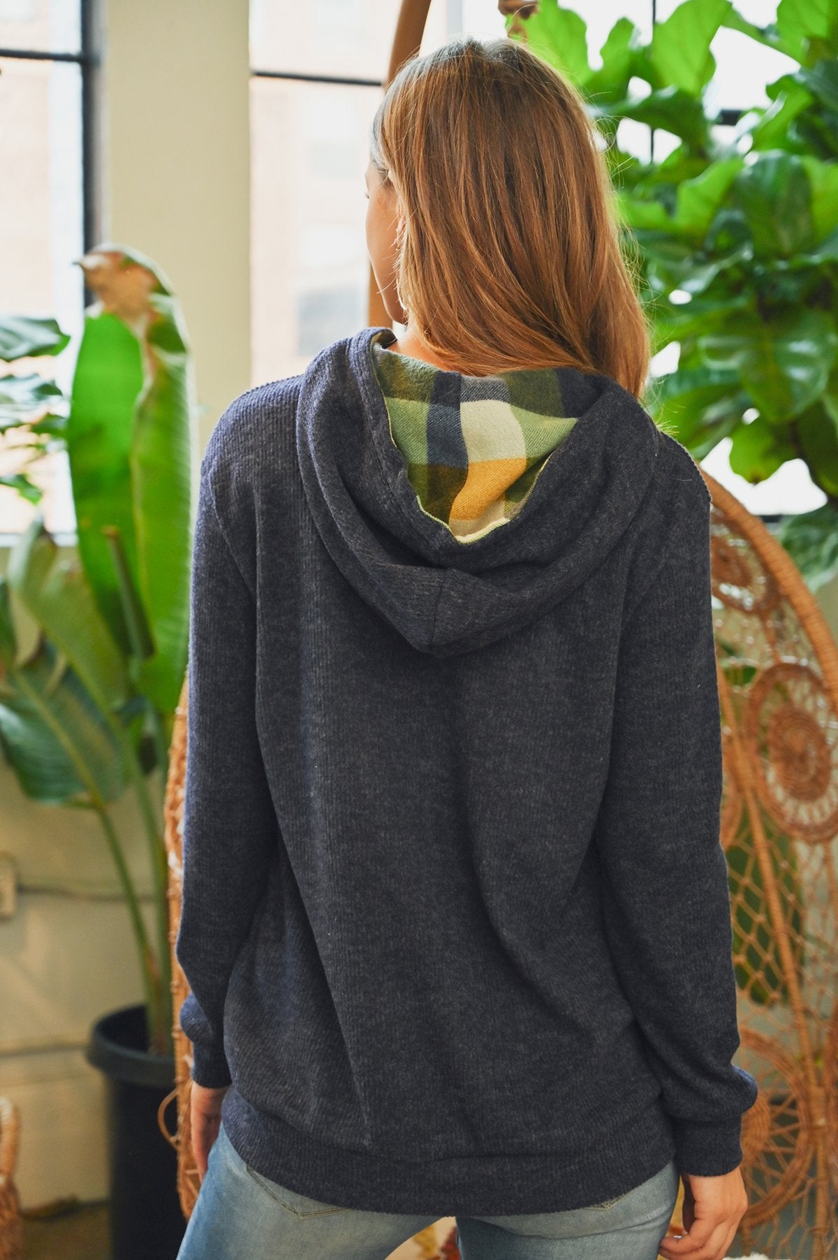 Someone wearing the Riah Fashion Hacci Brushed Plaid Kangaroo Pocket Hoodie stands indoors with plants in the background.