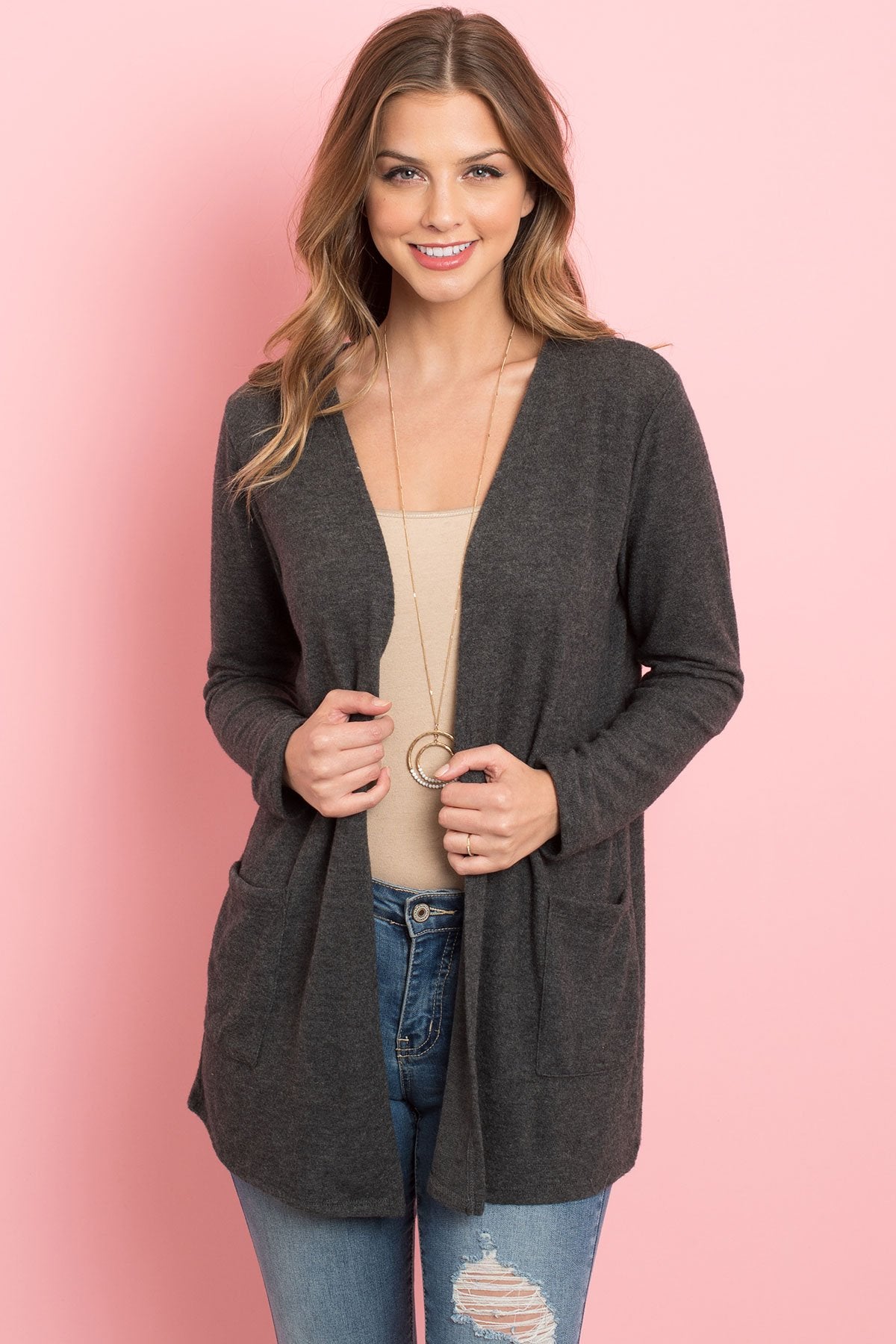 A woman in a teal Solid Long Sleeved Open Front Pocket Cardigan by Riah Fashion over a beige top stands against a pink background, smiling with hands in pockets.