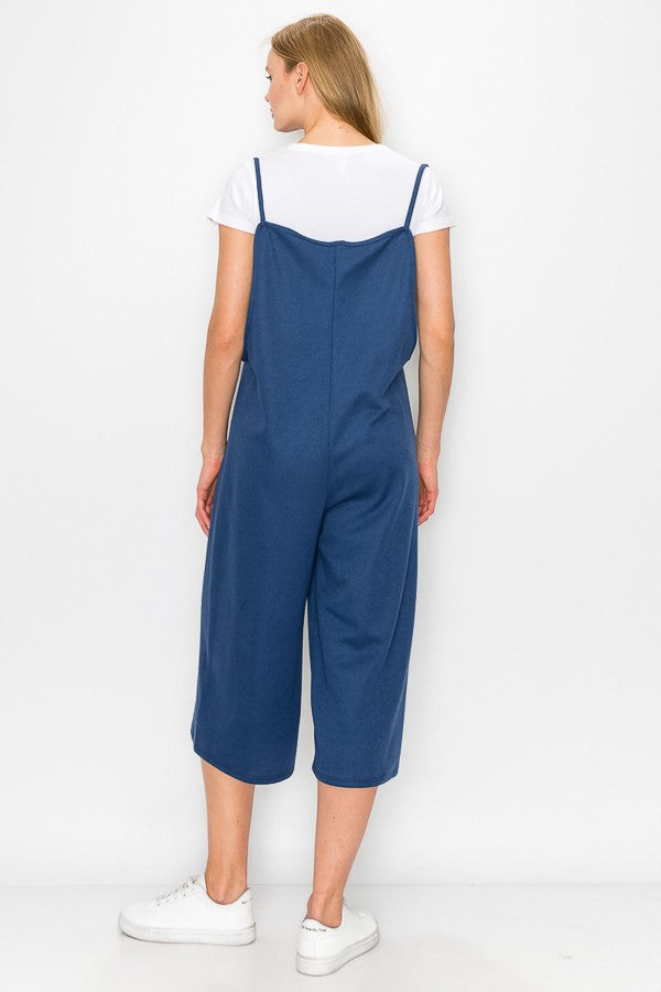 A person wearing a Stylespect Cropped Bottom Wide Leg Oversized Jumpsuit in blue over a white T-shirt and white sneakers, standing and smiling against a plain white background.