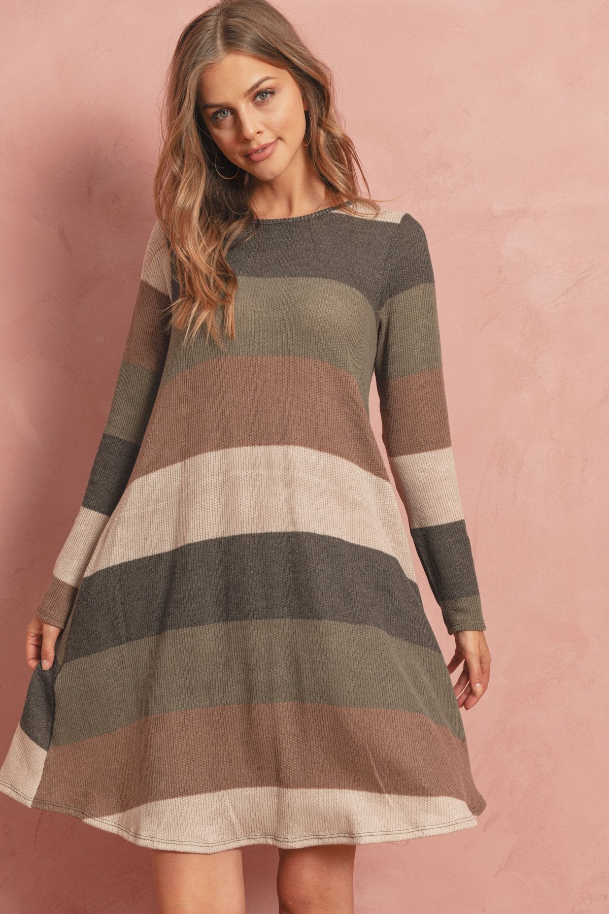 A woman poses in the Riah Fashion Long Sleeved Rib Stripe Pocket Dress in Charcoal/Mocha against a pink textured background.