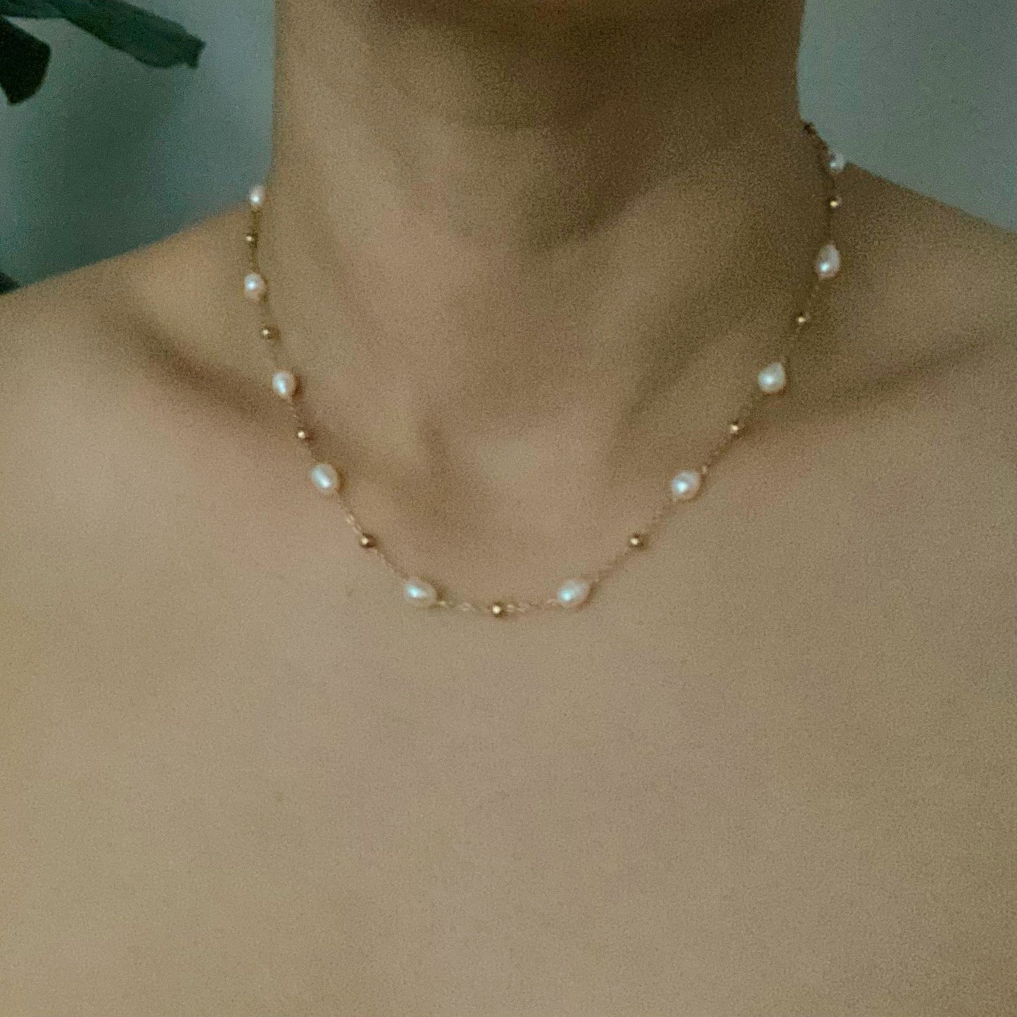 The Ashley Freshwater Pearl Necklace by Ellison + Young is a delicate gold chain adorned with small pearls and tiny gold beads, elegantly displayed on a light marble surface with green leaves and white stones.