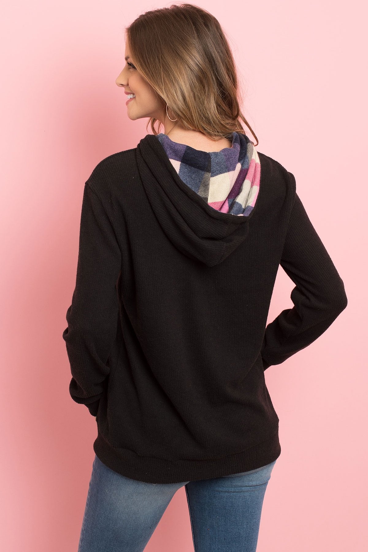 Someone wearing the Riah Fashion Hacci Brushed Plaid Kangaroo Pocket Hoodie stands indoors with plants in the background.