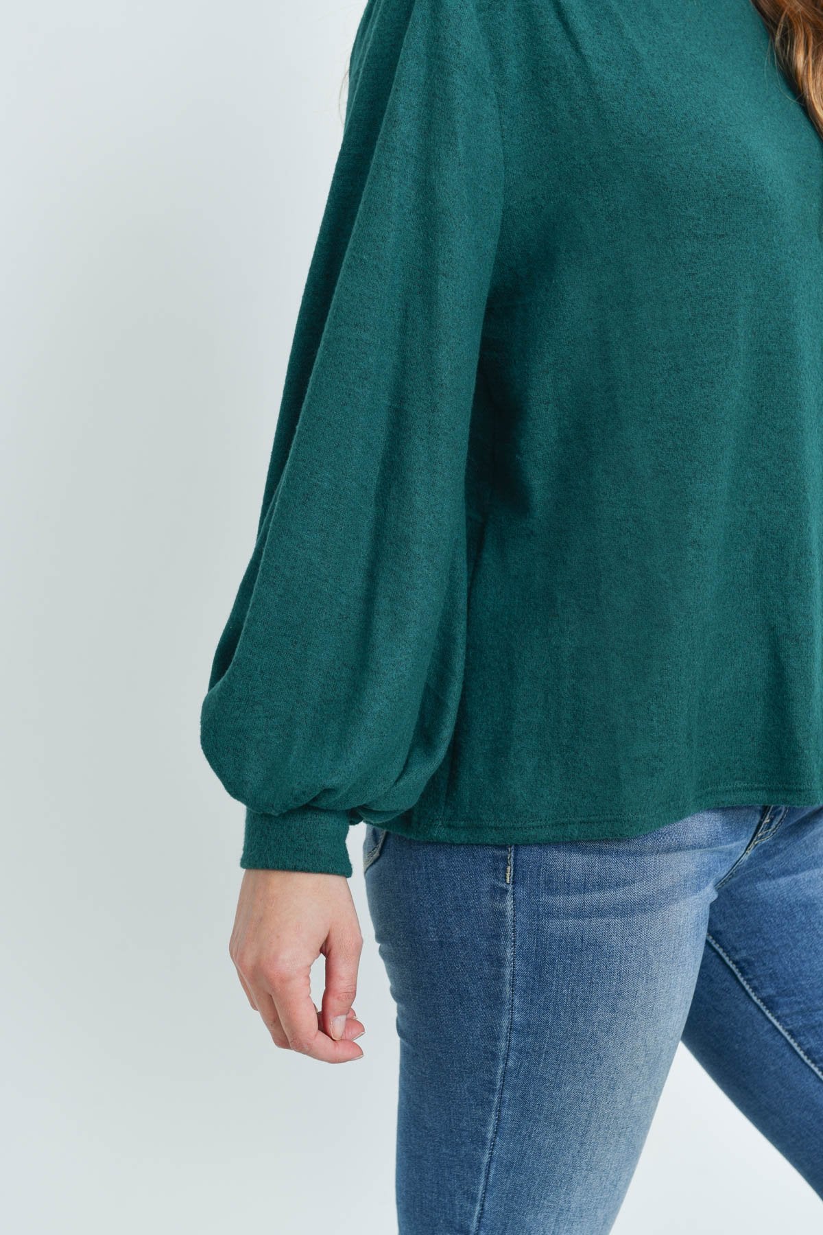 A woman models a Riah Fashion Puff Sleeved Boat Neck Two Toned Brushed Hacci Top with ripped jeans and brown boots on a concrete floor against a plain backdrop, accessorized with a long necklace.
