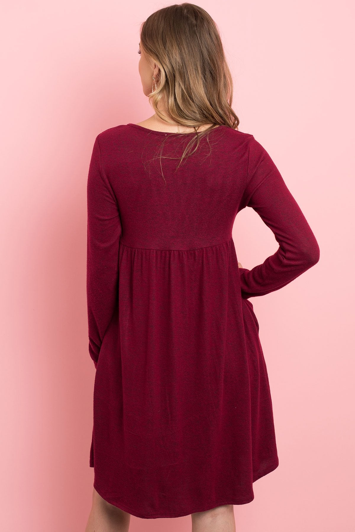 A woman in a Riah Fashion Boat Neck Brushed Hacci Round Hem Dress in Dark Ruby stands before a pink backdrop, adorned with a long necklace featuring circular pendants.