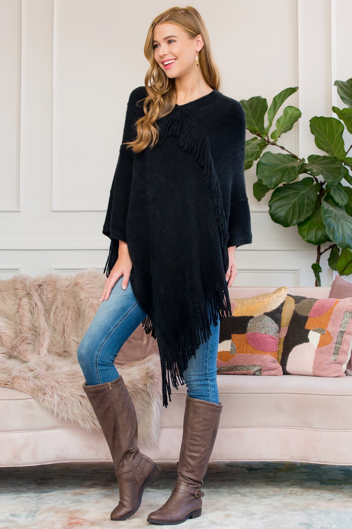 A person stands wearing a Riah Fashion Asymmetric Fringe Poncho in one color, paired with jeans and brown boots, smiling while holding the poncho. In the background are a plant and a sofa with cushions.