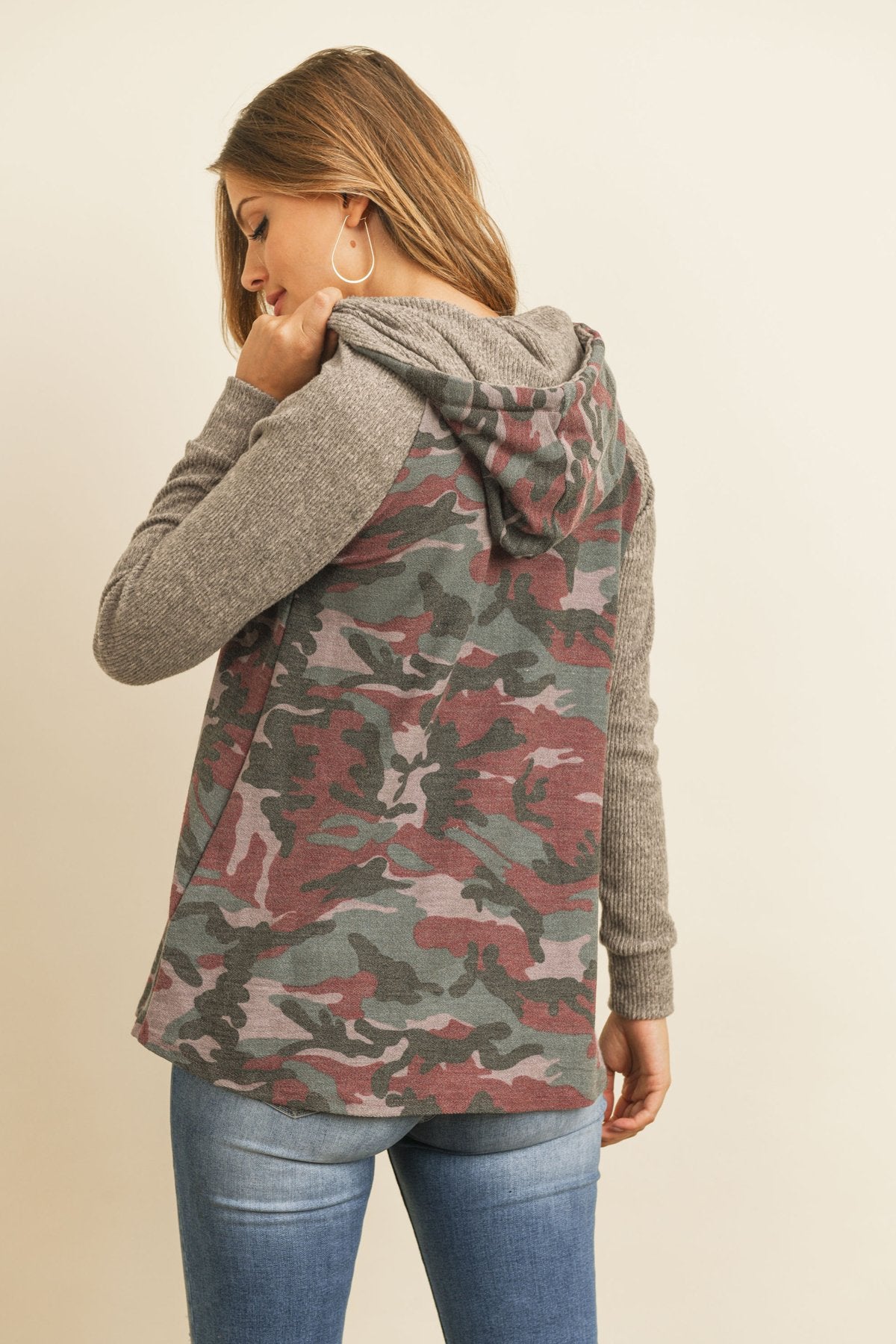 A person wearing the Self Tie Rib Hacci Brushed Sleeve Camouflage Hoodie by Riah Fashion with distressed jeans, holding the hoodie strings, stands against a beige background.