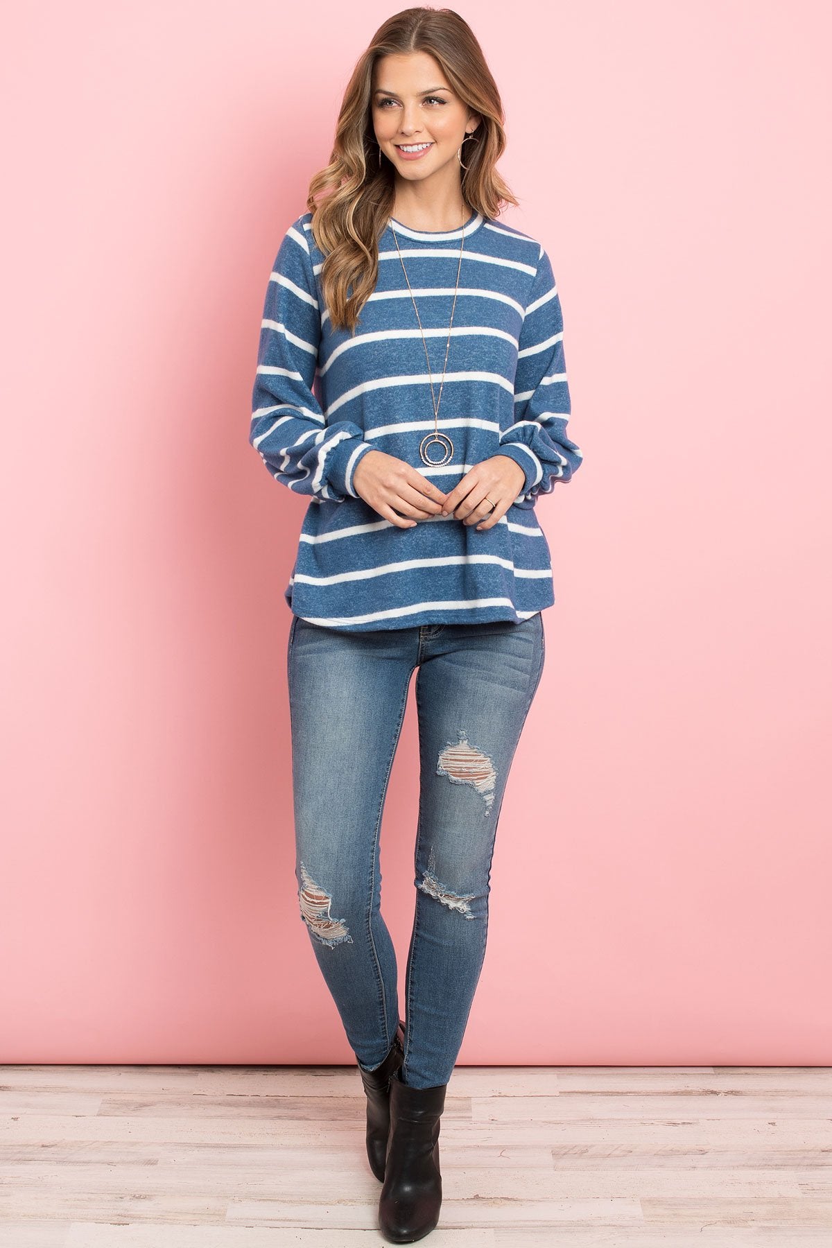 A woman in a Riah Fashion Striped Long Sleeved Round Neck Swing Top and jeans stands against a pink backdrop, wearing a long necklace and touching her hair.