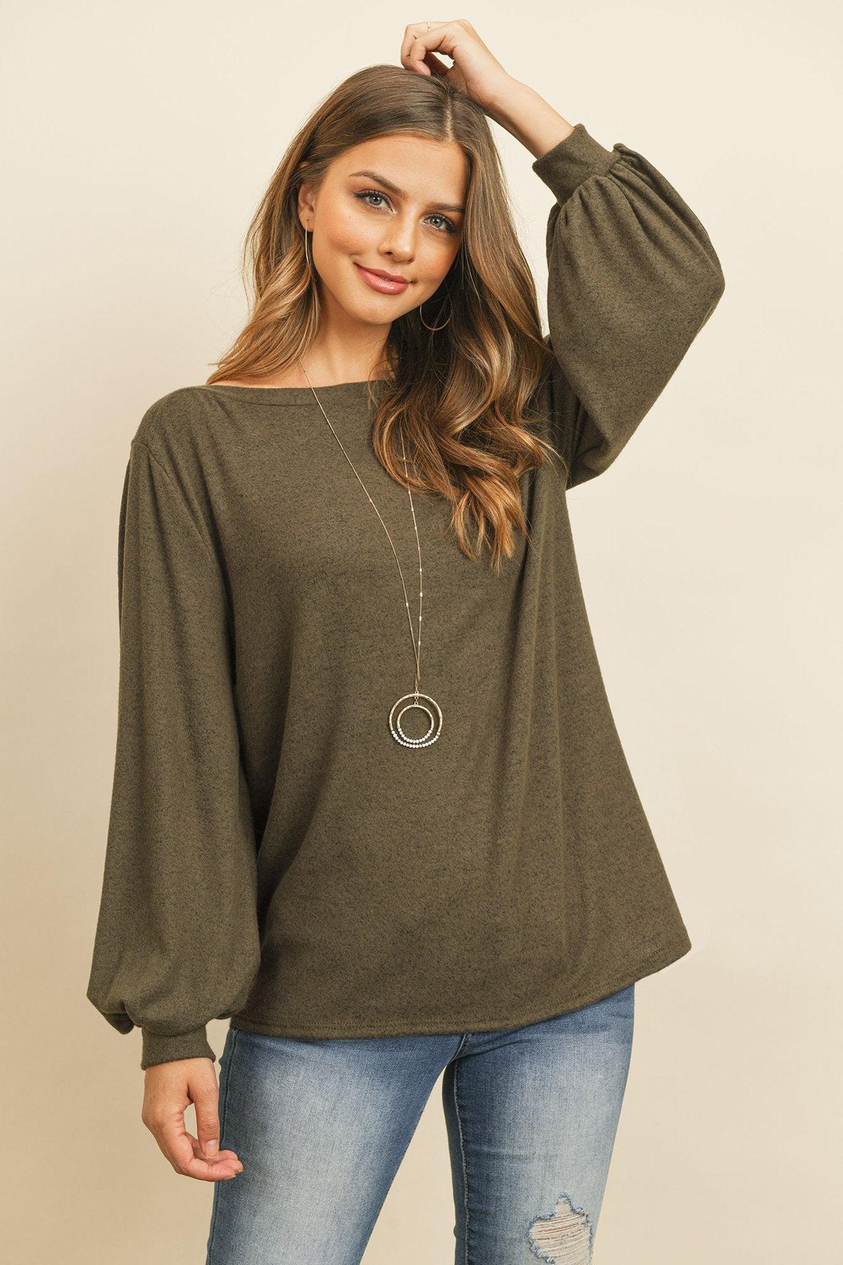 A woman models a Riah Fashion Puff Sleeved Boat Neck Two Toned Brushed Hacci Top with ripped jeans and brown boots on a concrete floor against a plain backdrop, accessorized with a long necklace.