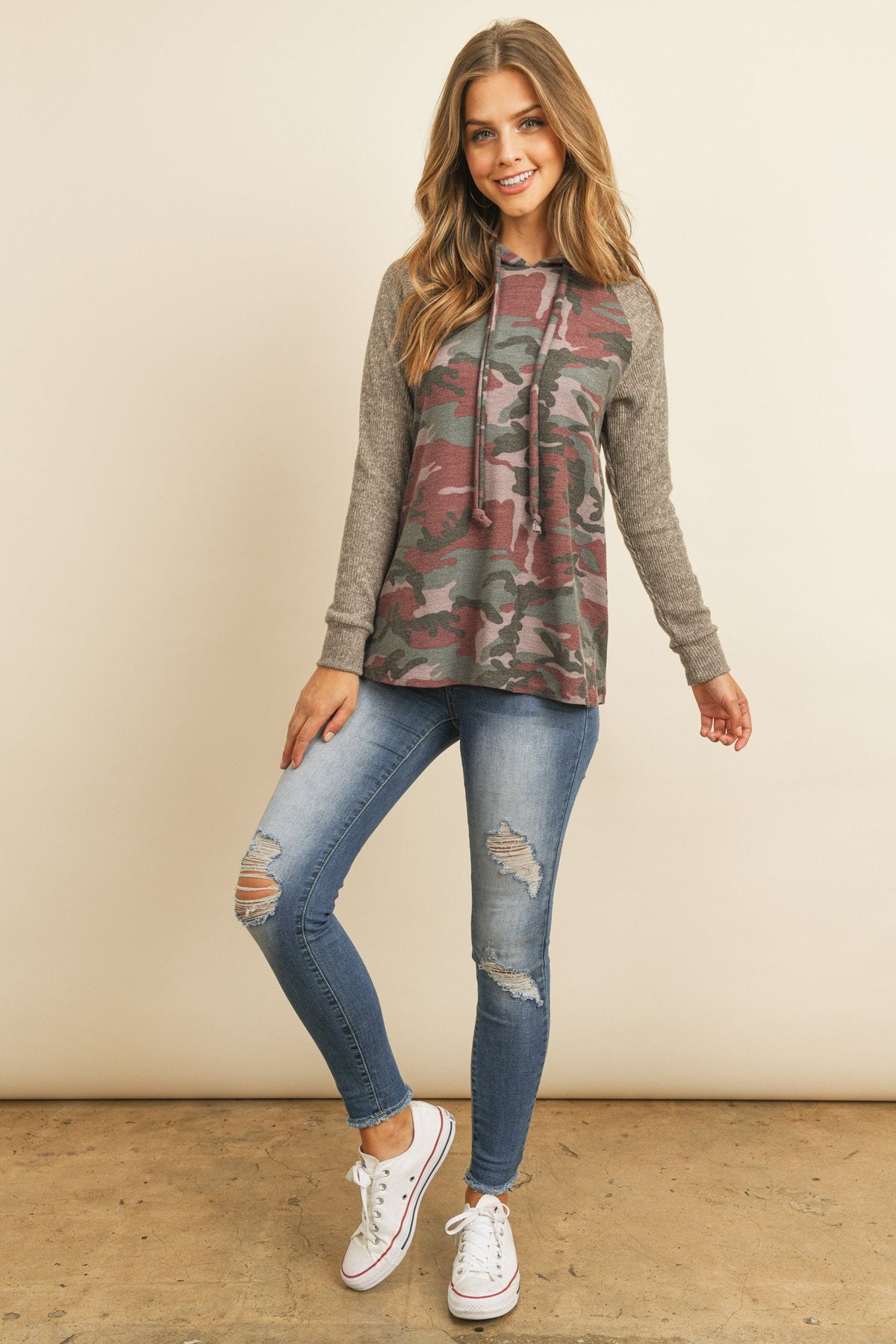 A person wearing the Self Tie Rib Hacci Brushed Sleeve Camouflage Hoodie by Riah Fashion with distressed jeans, holding the hoodie strings, stands against a beige background.