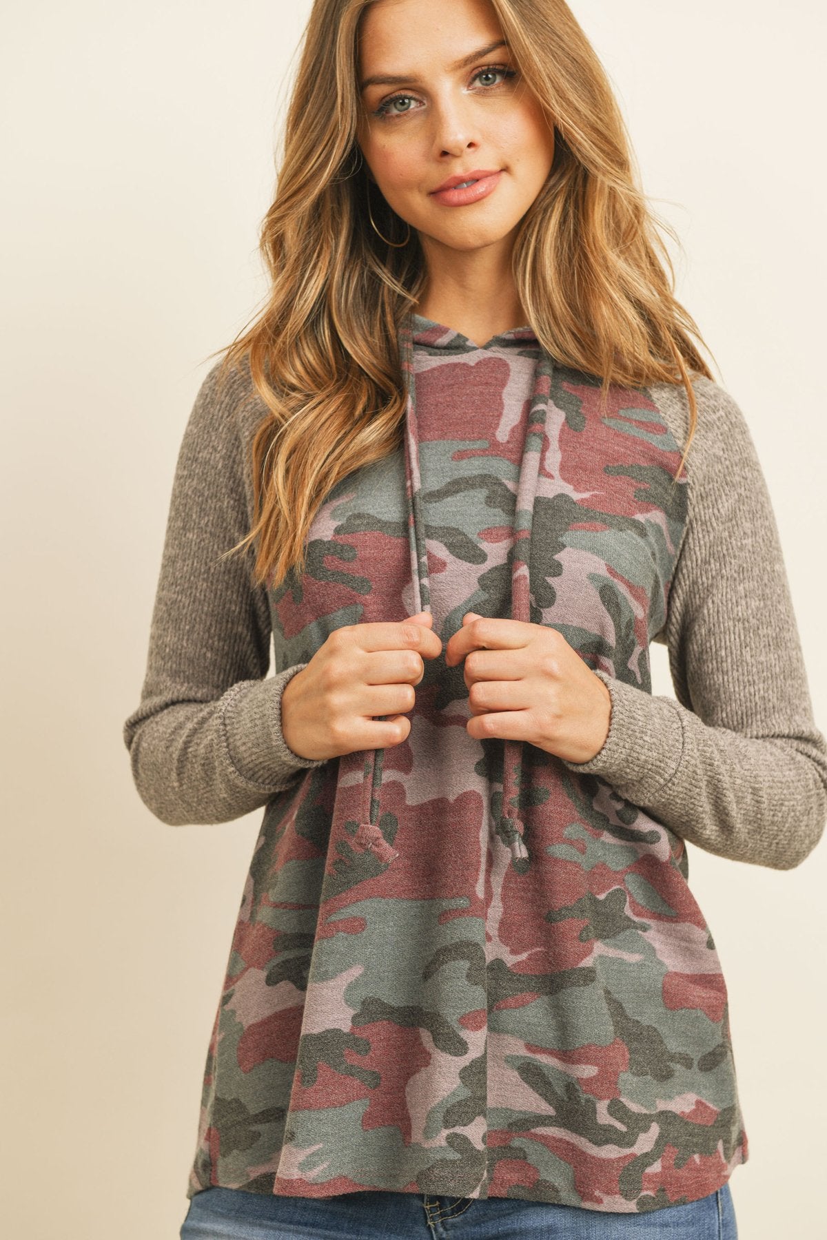 A person wearing the Self Tie Rib Hacci Brushed Sleeve Camouflage Hoodie by Riah Fashion with distressed jeans, holding the hoodie strings, stands against a beige background.