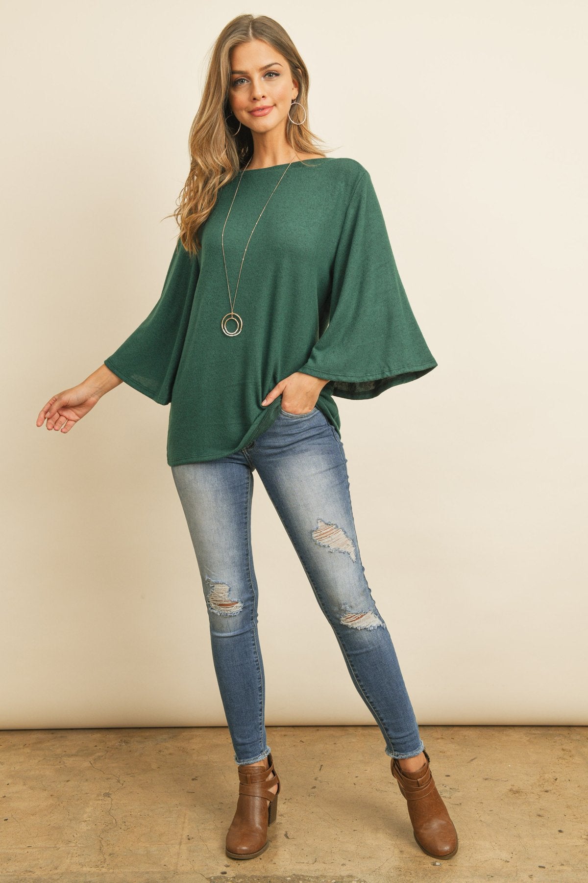 A person in a Riah Fashion Boat Neck Bell Sleeve Solid Hacci Brushed Top and ripped jeans stands against a neutral background, accessorized with hoop earrings and a long necklace.