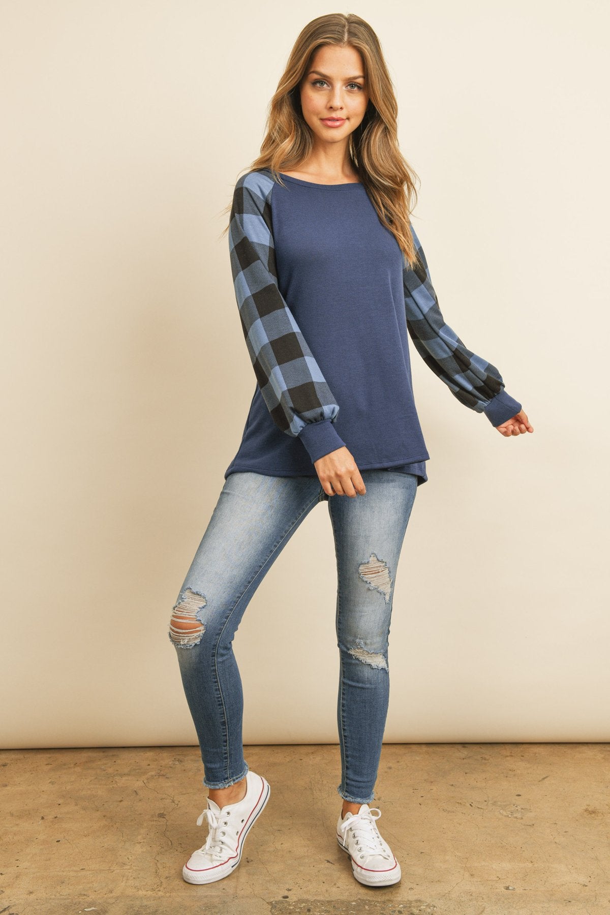 A woman wearing a faded denim plaid puff sleeved round neck top by Riah Fashion and ripped jeans stands against a plain background.