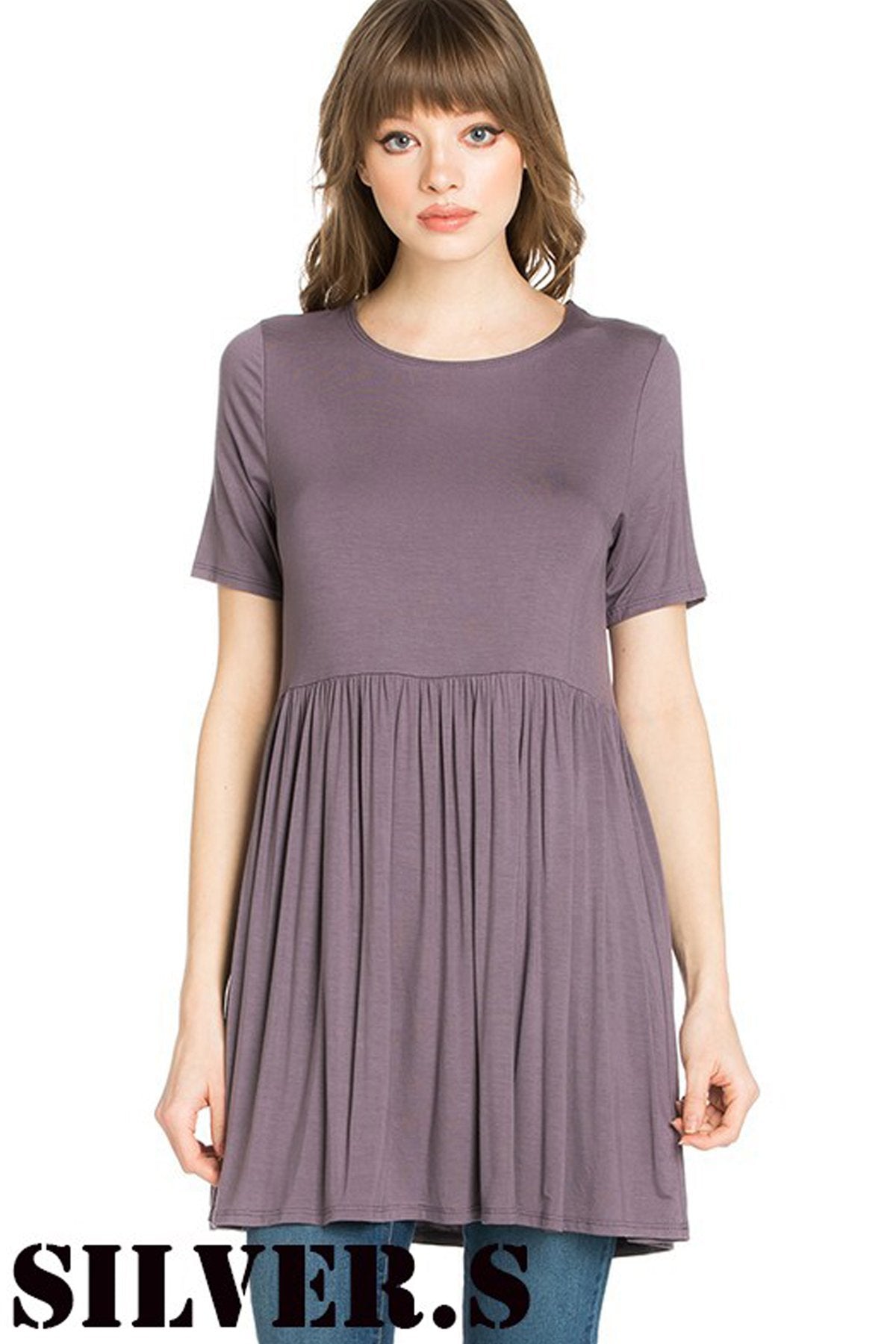 A woman stands outdoors wearing a beige Riah Fashion Short Sleeve Empire Waist Tunic paired with a long necklace.