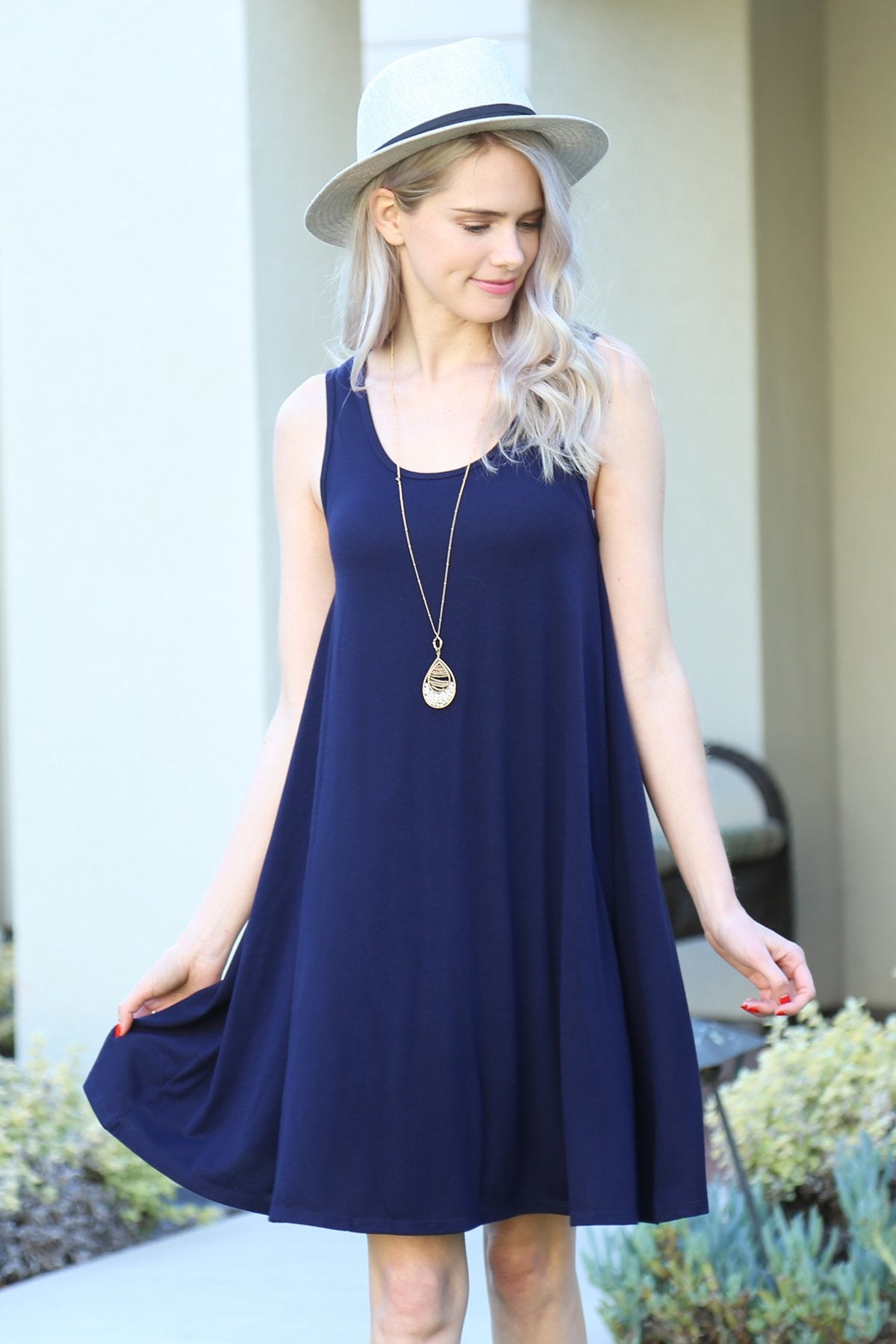 A person with long, light-colored hair wears a Sleeveless Flared Pocket Dress in light green by Riah Fashion outside, holding the dresss sides along with a triangular pendant necklace.