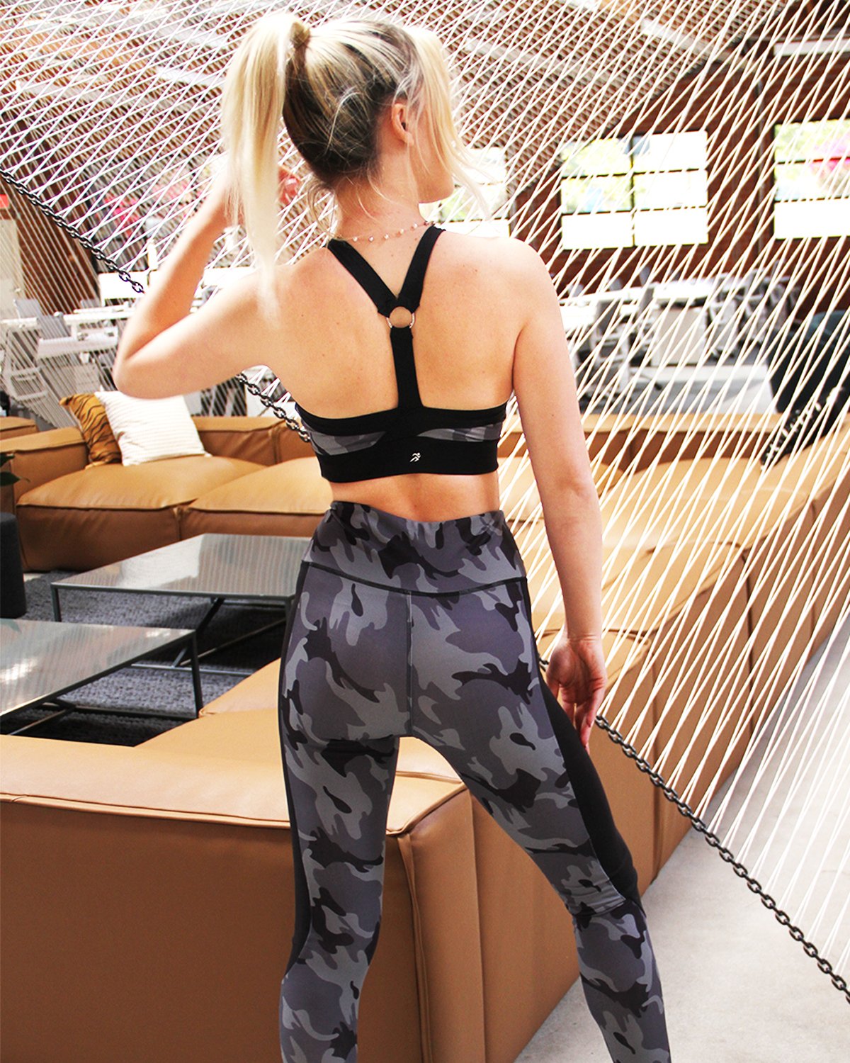 Wearing the Veloso Supplex Moisture-Resistant Fashion Leggings & Sports Bra Set by Savoy Active, a person in camo-patterned workout attire poses indoors with one hand on a swinging chair.