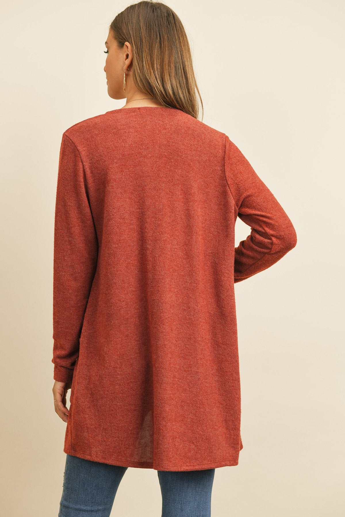 A woman in a teal Solid Long Sleeved Open Front Pocket Cardigan by Riah Fashion over a beige top stands against a pink background, smiling with hands in pockets.