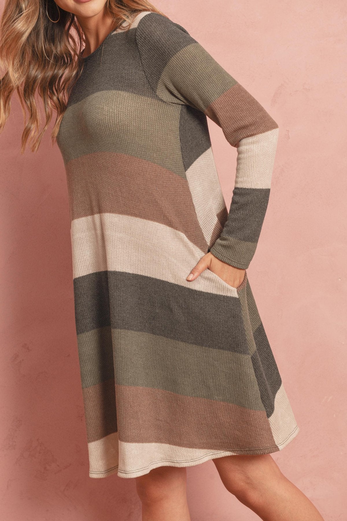 A woman poses in the Riah Fashion Long Sleeved Rib Stripe Pocket Dress in Charcoal/Mocha against a pink textured background.