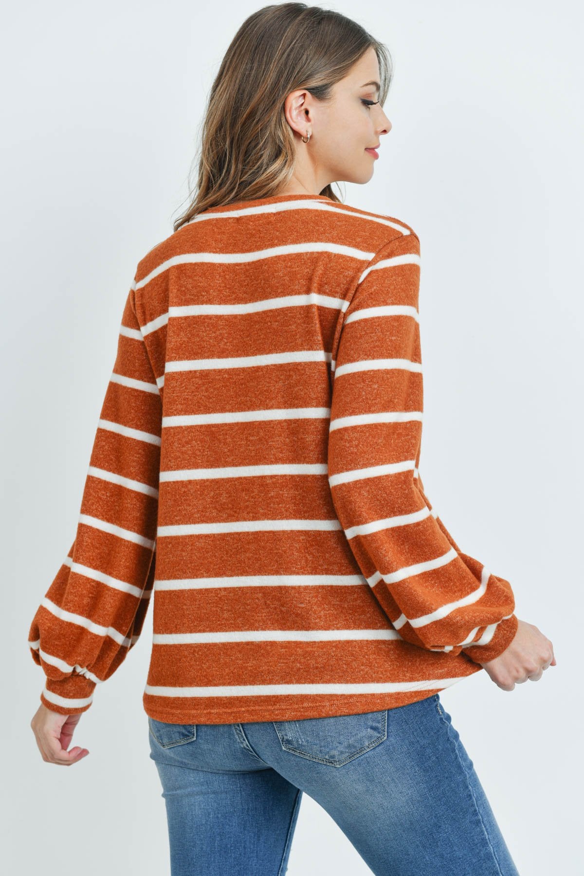 A woman in a Riah Fashion Striped Long Sleeved Round Neck Swing Top and jeans stands against a pink backdrop, wearing a long necklace and touching her hair.