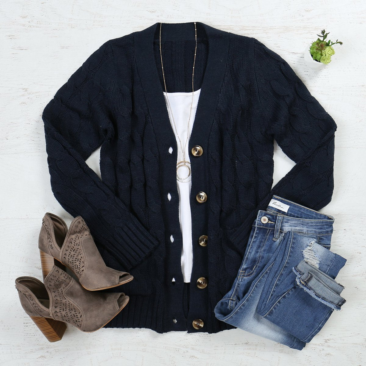 A person wearing an ash copper cable sweater with pockets from Riah Fashion stands in front of tall green plants and a wooden fence.
