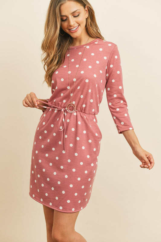 A woman smiles, wearing a Riah Fashion mauve/ivory Polka Dot Print French Terry Cinch Waist Tie Front Dress with pockets and a long necklace.