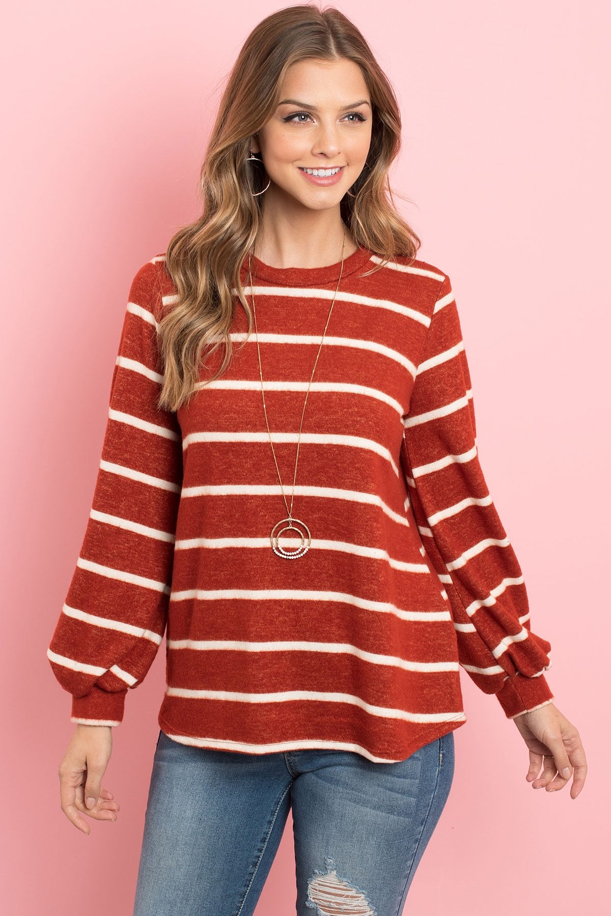 A woman in a Riah Fashion Striped Long Sleeved Round Neck Swing Top and jeans stands against a pink backdrop, wearing a long necklace and touching her hair.