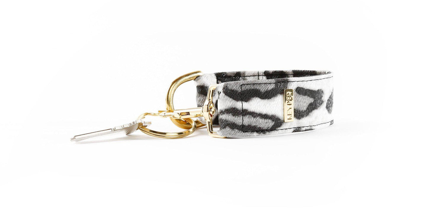 The KEYPER brands Snow Leopard Vegan Leather KEYPER® Key Ring features a leopard print design, a gold key ring with an attached key, and a small rectangular gold label.