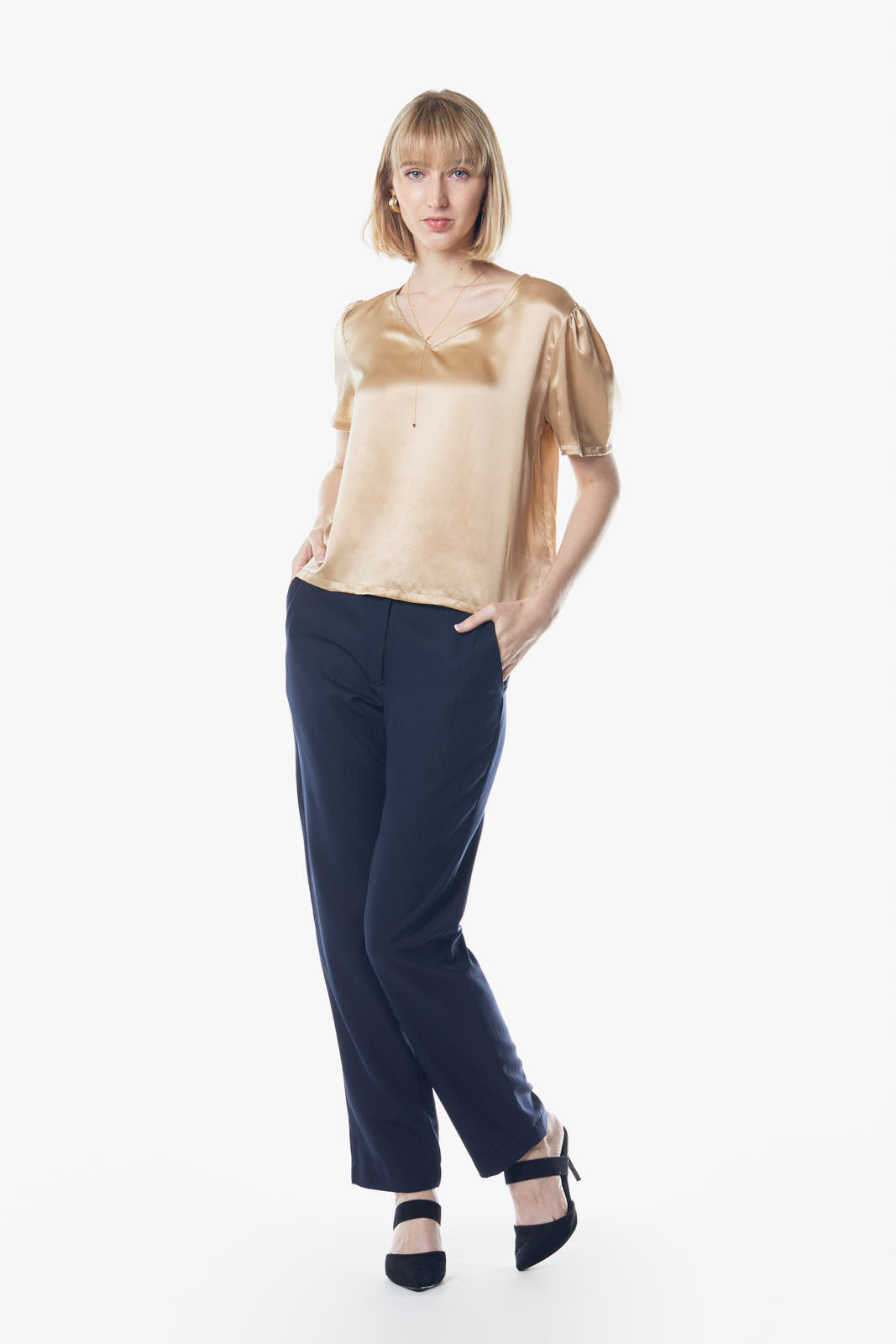 A person wearing the Le Réussi® Champagne V-Neck Blouse and dark pants stands against a white background, hands in pockets.