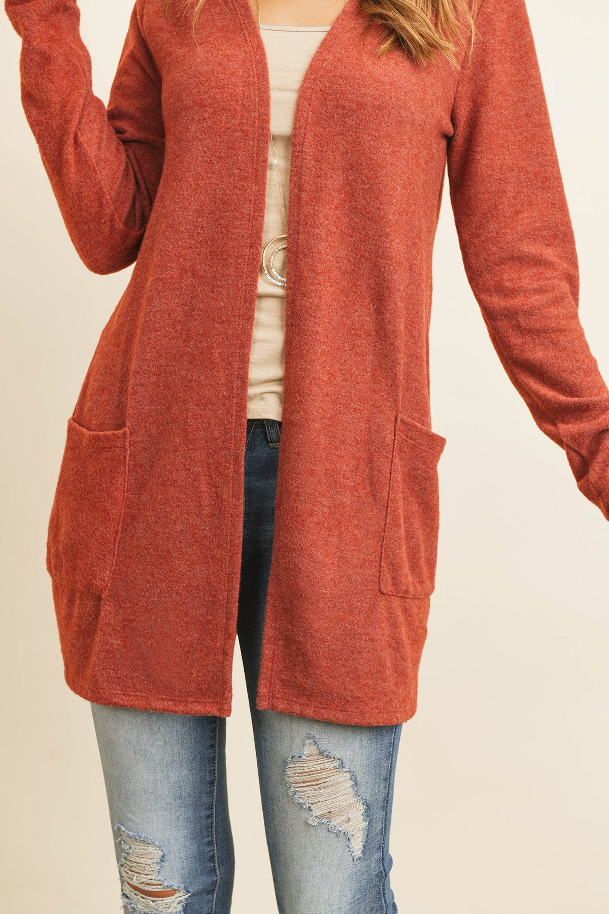 A woman in a teal Solid Long Sleeved Open Front Pocket Cardigan by Riah Fashion over a beige top stands against a pink background, smiling with hands in pockets.