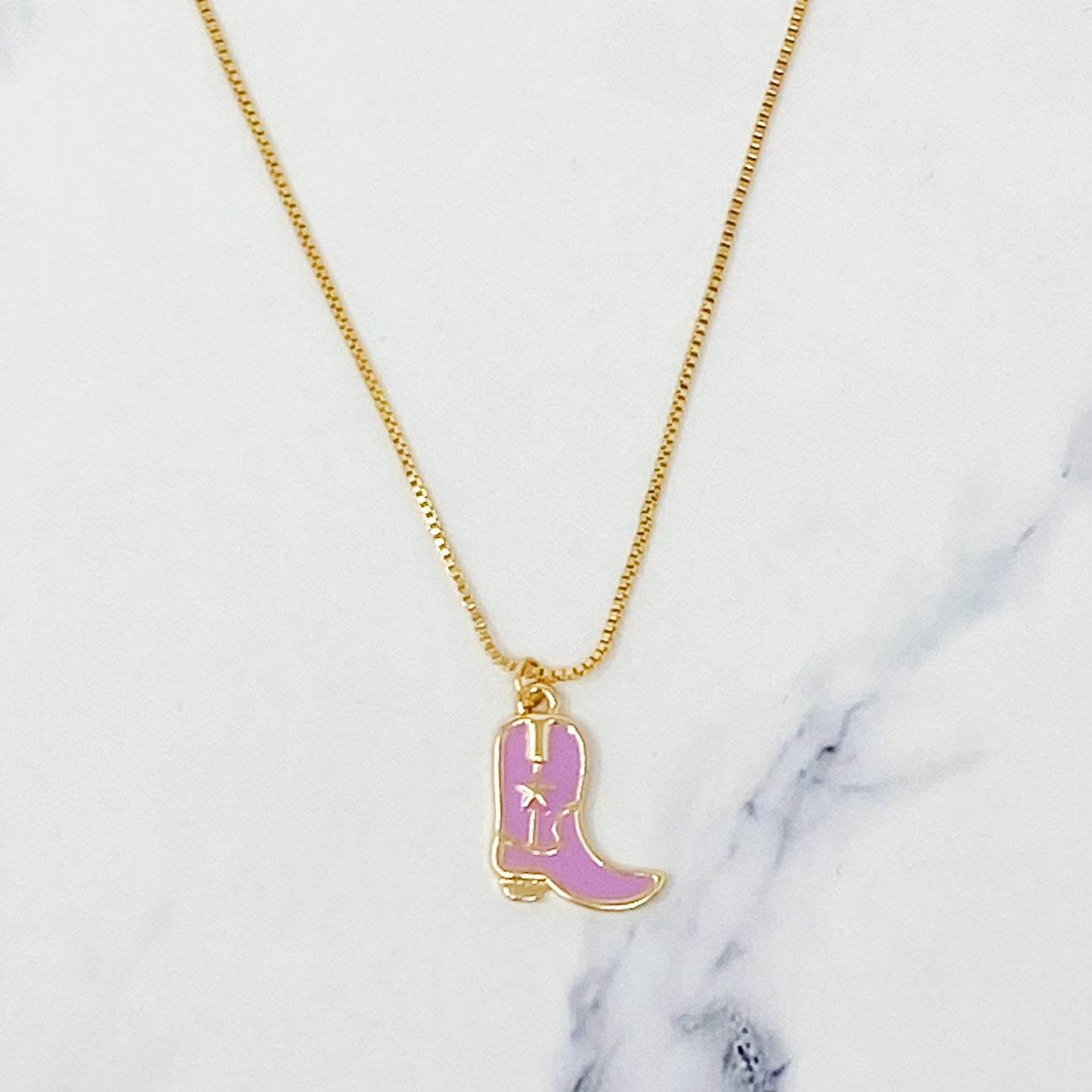 Two Cow Girl Boot Pendant Necklaces by Ellison + Young, one with a white boot and the other with a pink boot, are displayed on a marble surface.