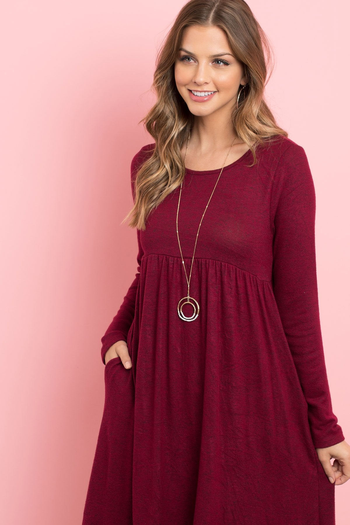 A woman in a Riah Fashion Boat Neck Brushed Hacci Round Hem Dress in Dark Ruby stands before a pink backdrop, adorned with a long necklace featuring circular pendants.
