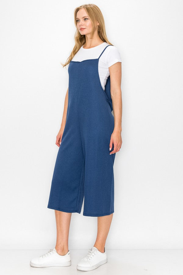 A person wearing a Stylespect Cropped Bottom Wide Leg Oversized Jumpsuit in blue over a white T-shirt and white sneakers, standing and smiling against a plain white background.