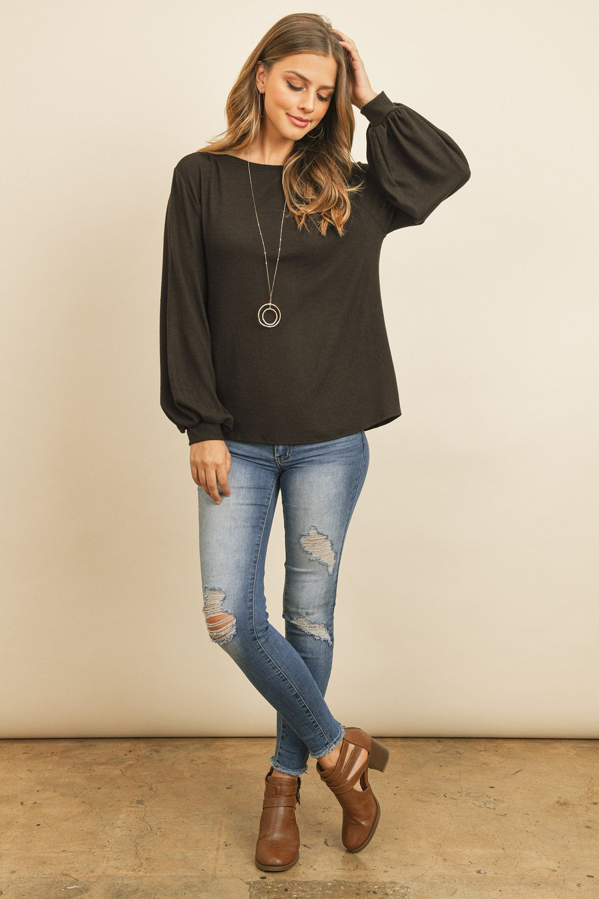 A woman models a Riah Fashion Puff Sleeved Boat Neck Two Toned Brushed Hacci Top with ripped jeans and brown boots on a concrete floor against a plain backdrop, accessorized with a long necklace.