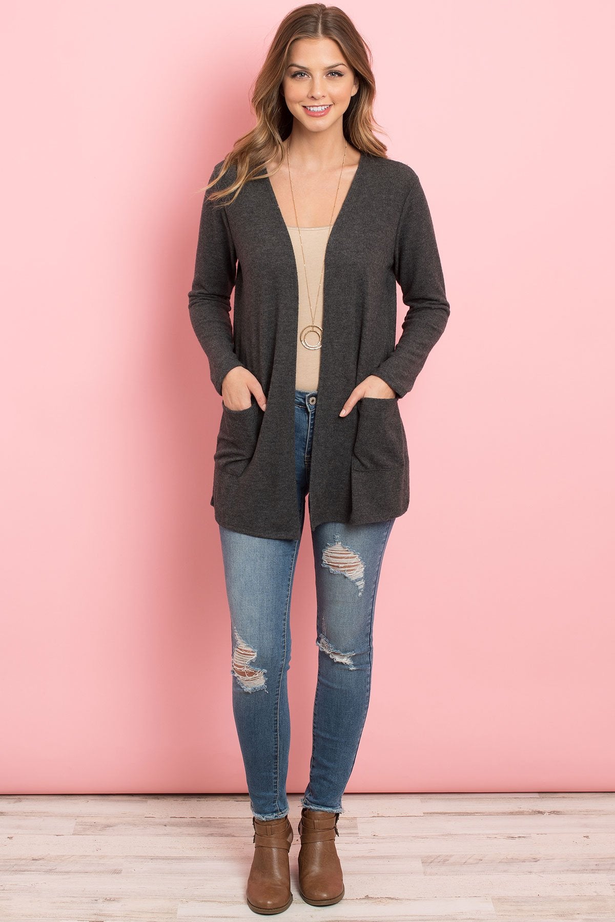 A woman in a teal Solid Long Sleeved Open Front Pocket Cardigan by Riah Fashion over a beige top stands against a pink background, smiling with hands in pockets.