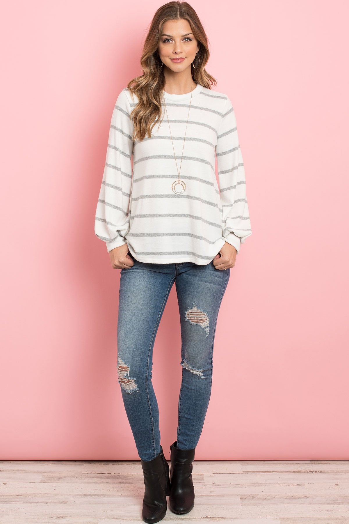 A woman in a Riah Fashion Striped Long Sleeved Round Neck Swing Top and jeans stands against a pink backdrop, wearing a long necklace and touching her hair.