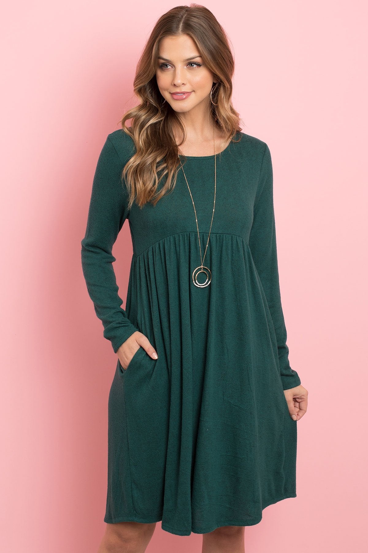 A woman in a Riah Fashion Boat Neck Brushed Hacci Round Hem Dress in Dark Ruby stands before a pink backdrop, adorned with a long necklace featuring circular pendants.