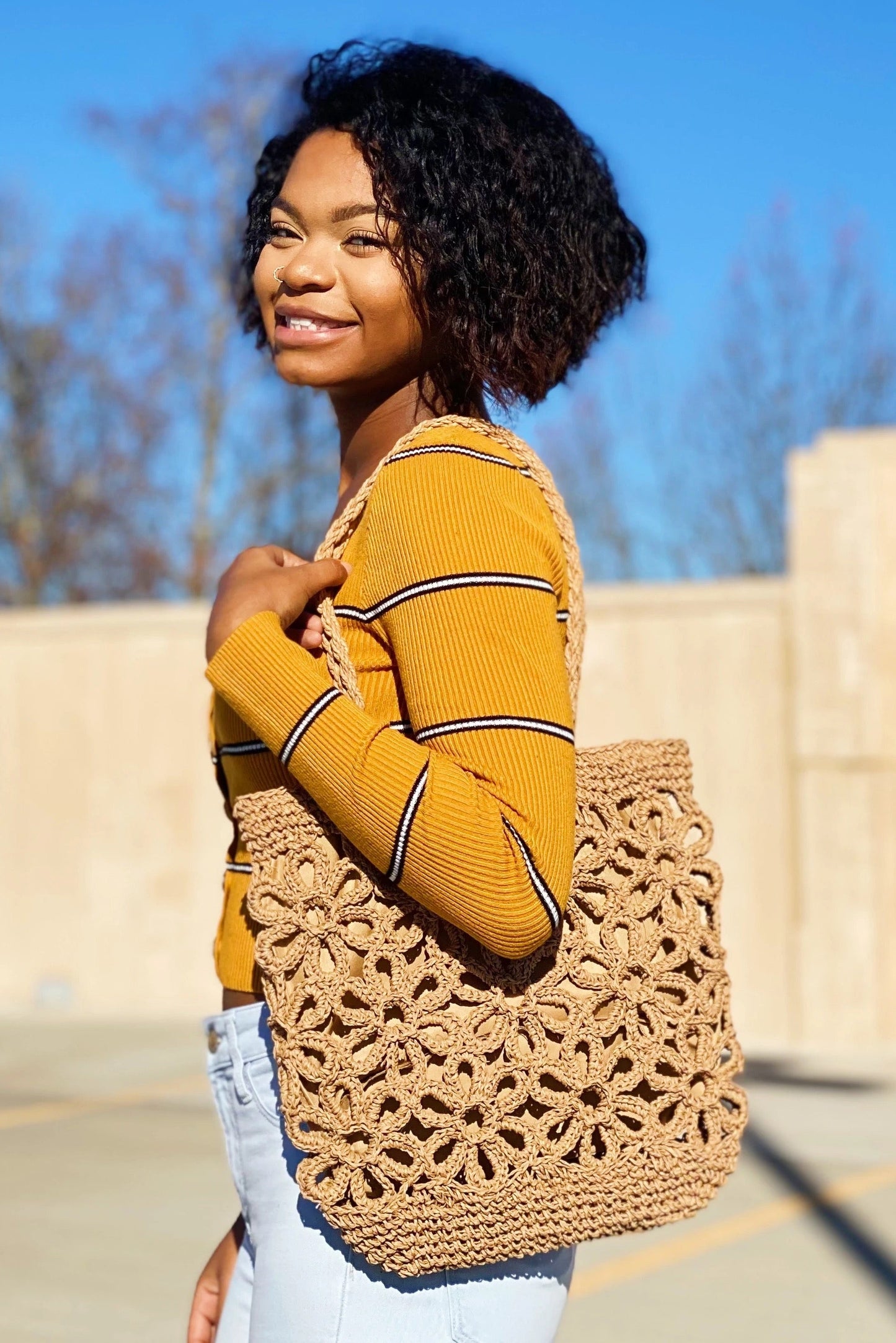 A person smiles while standing outdoors with a clear blue sky and trees in the background, wearing a yellow striped sweater and holding an Ellison + Young Hand Made Muted Floral Tote.