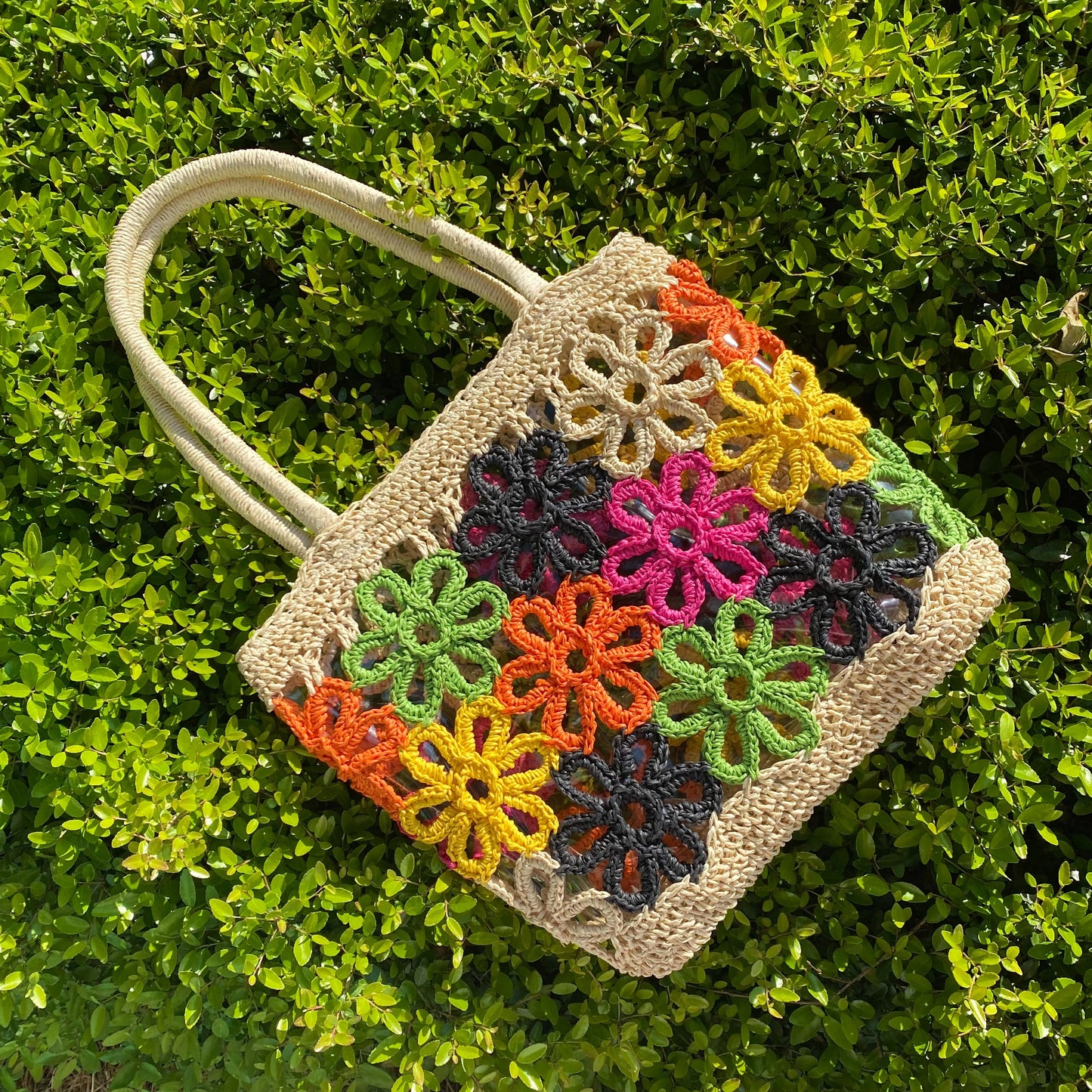 The Hand Made Flower Power Tote by Ellison + Young, featuring colorful floral crochet patterns in orange, green, pink, black, and yellow, rests on dense green foliage.