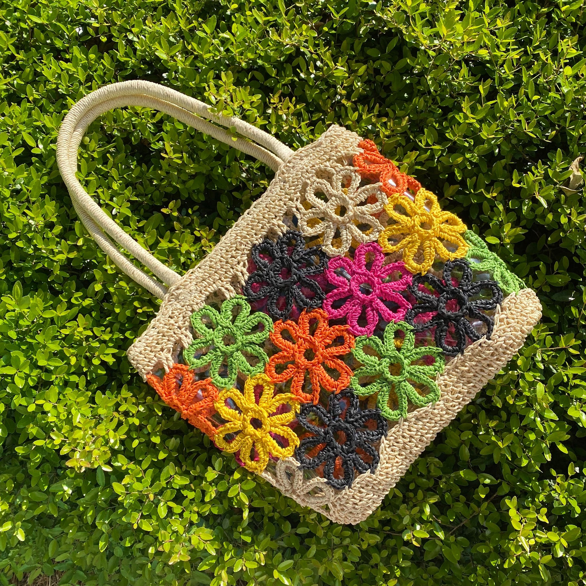 The Hand Made Flower Power Tote by Ellison + Young, featuring colorful floral crochet patterns in orange, green, pink, black, and yellow, rests on dense green foliage.