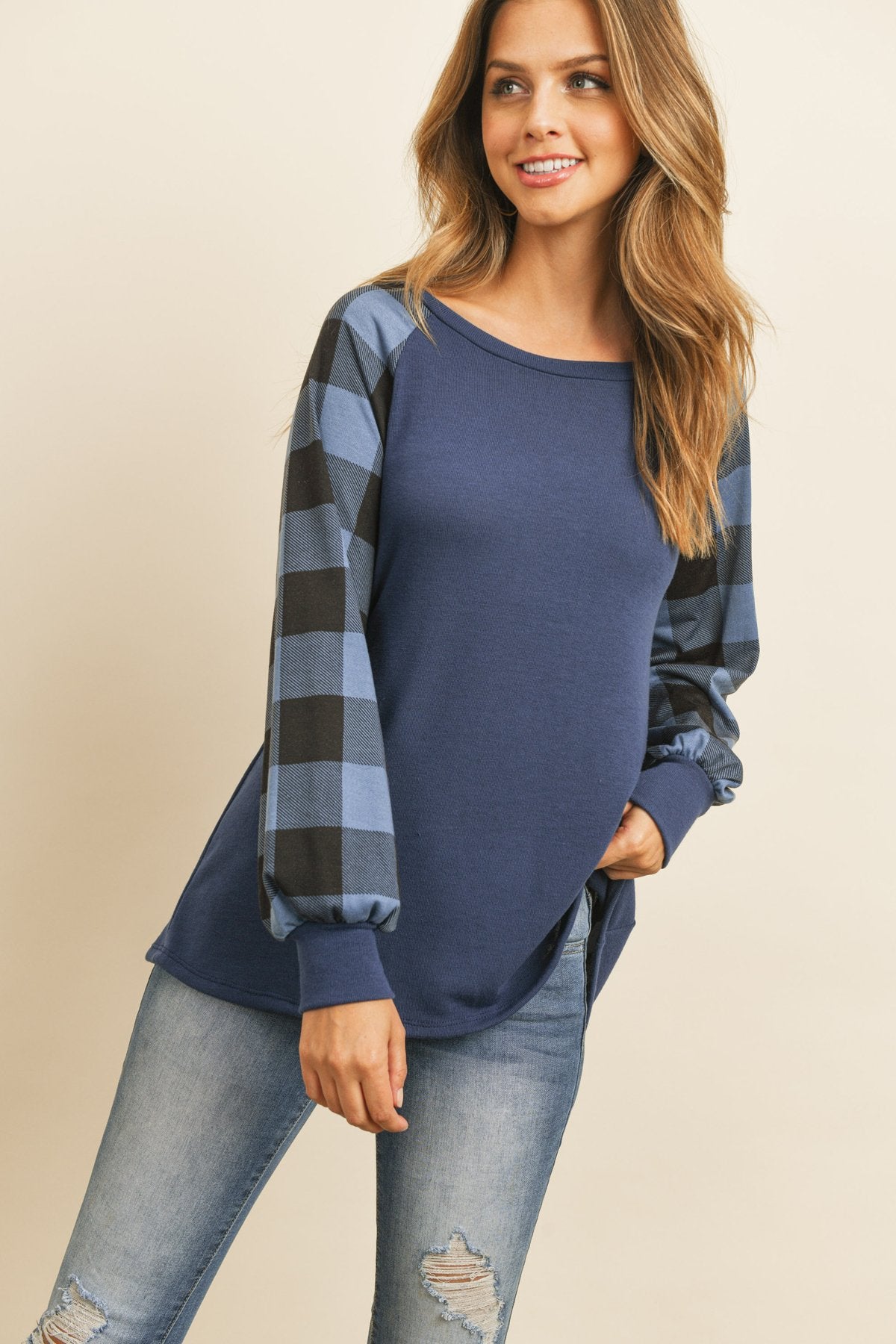A woman wearing a faded denim plaid puff sleeved round neck top by Riah Fashion and ripped jeans stands against a plain background.